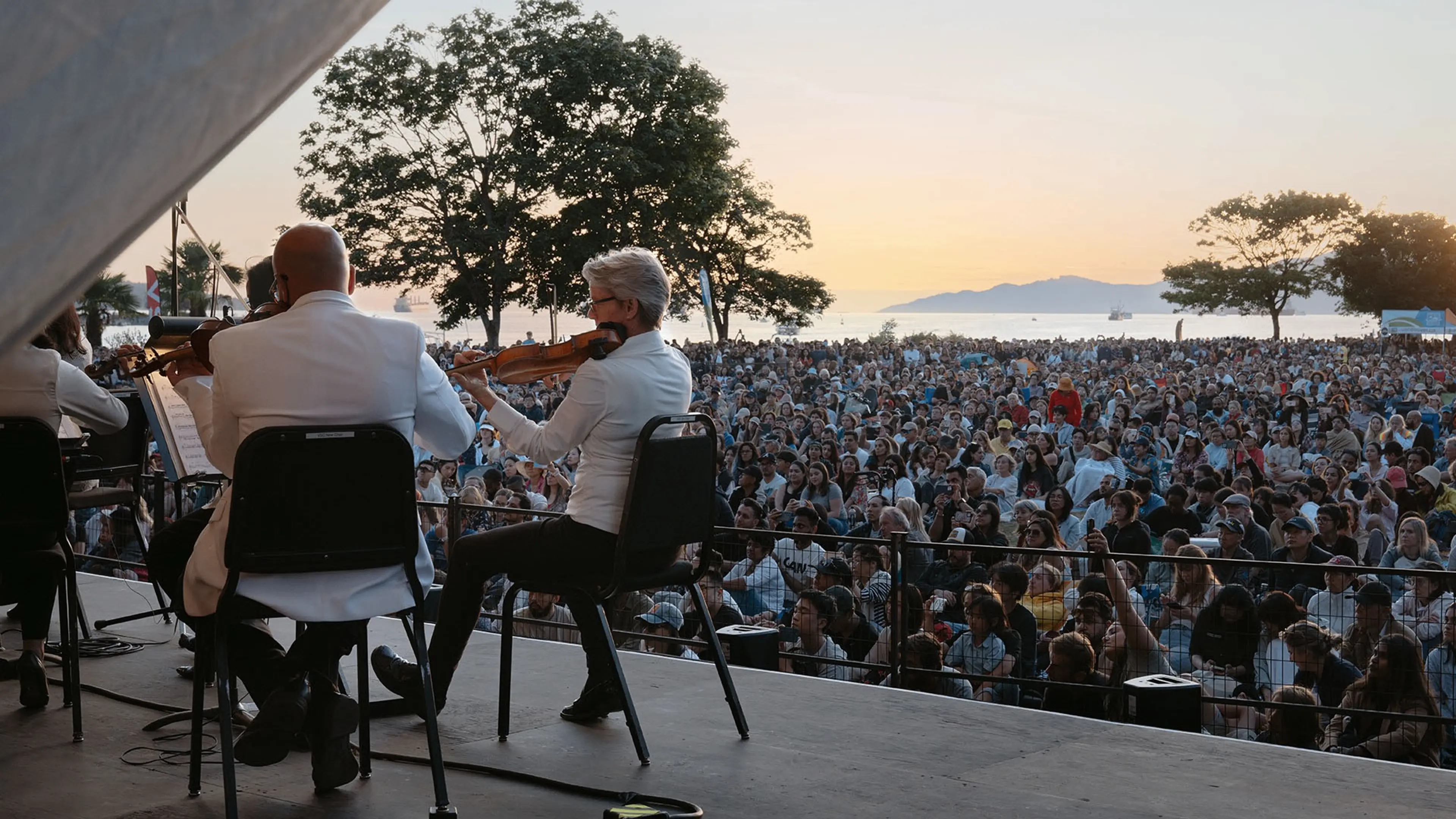 Musicians in white jackets playing violins on stage facing a large outdoor audience at sunset near water and trees.