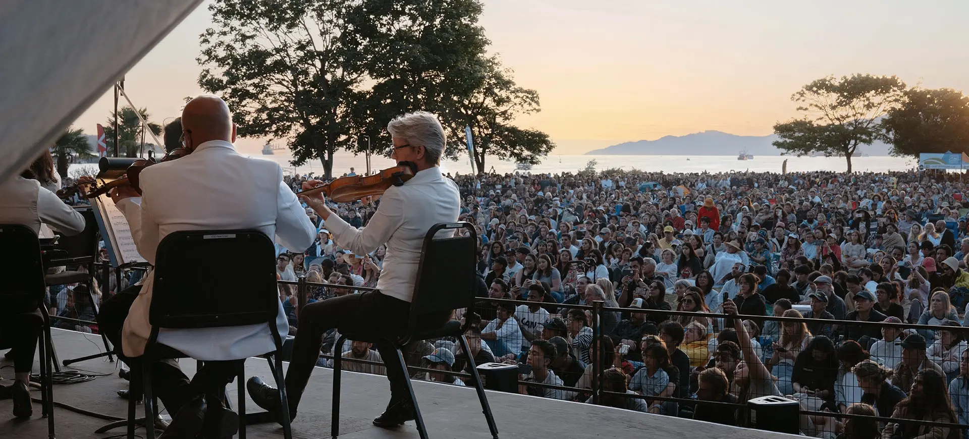Musicians in white jackets playing violins on stage facing a large outdoor audience at sunset near water and trees.
