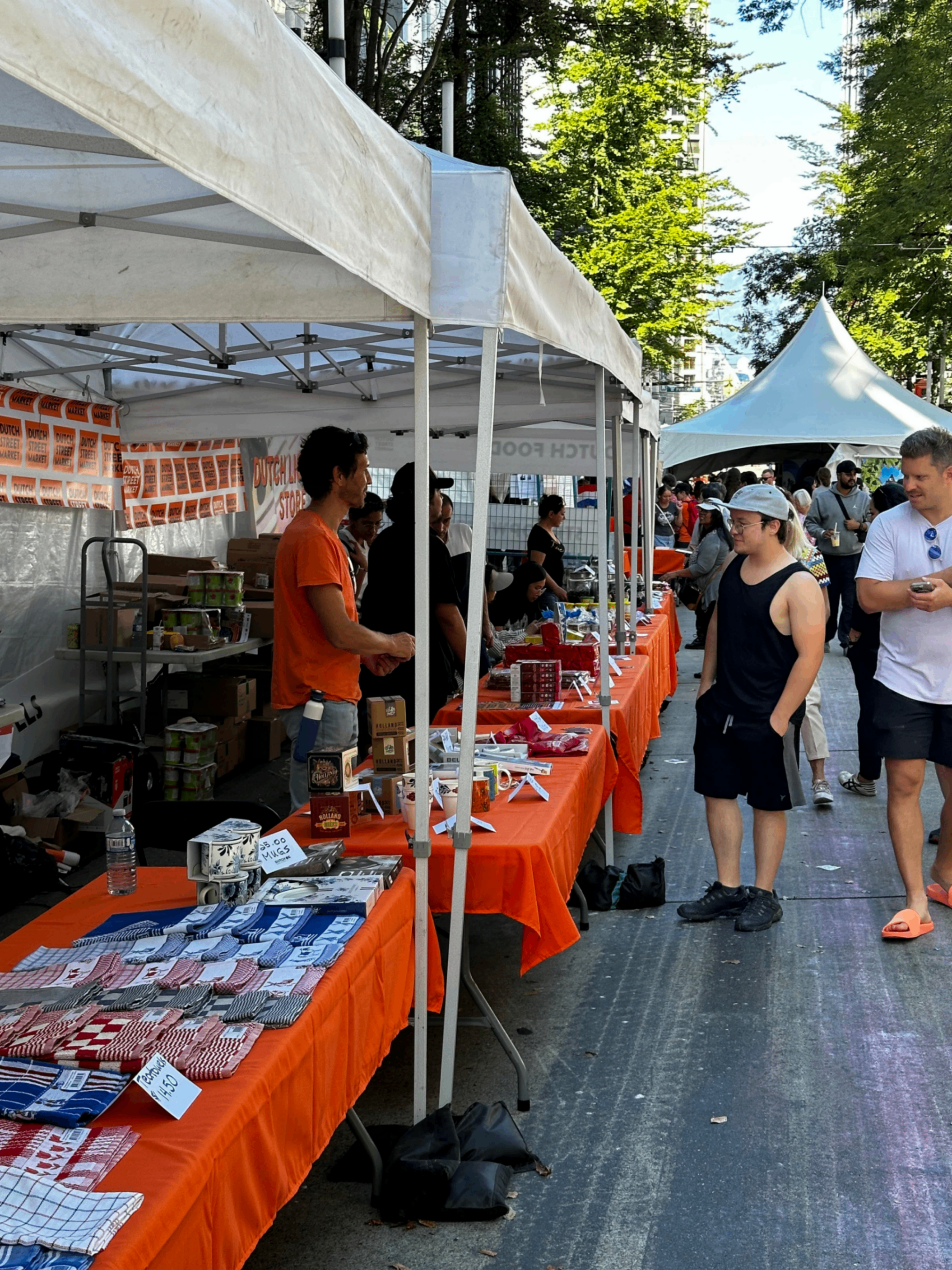 Outdoor market with white tents and orange tablecloths, people browsing and selling various goods on tables.