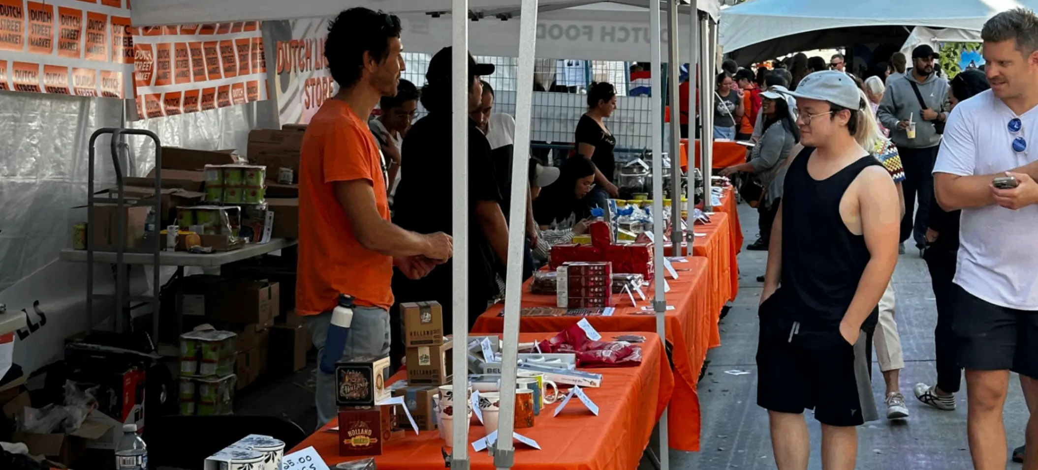 Outdoor market with white tents and orange tablecloths, people browsing and selling various goods on tables.