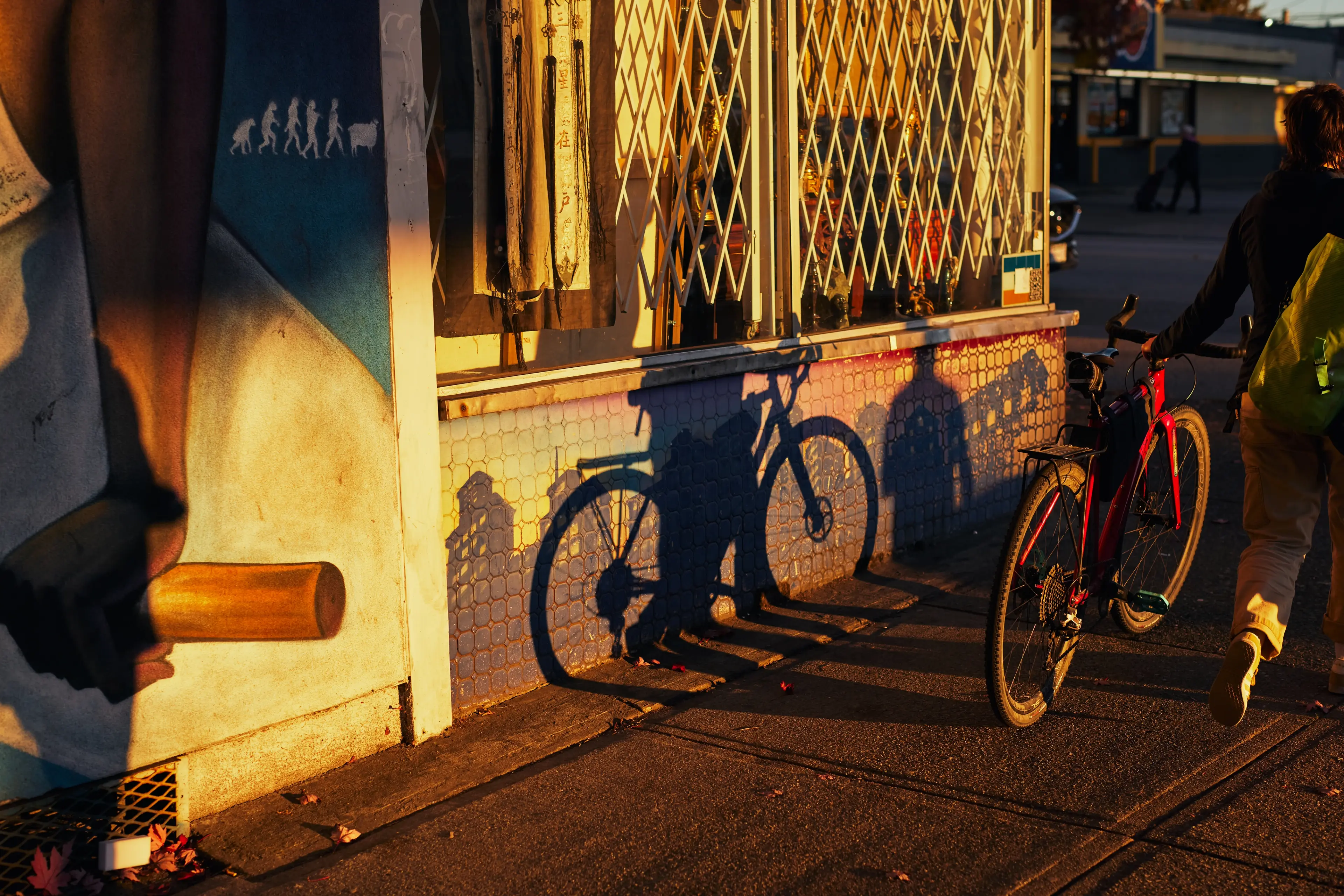 Person with a green backpack walking a red bicycle past a mural and casting long shadows on a wall at sunset.