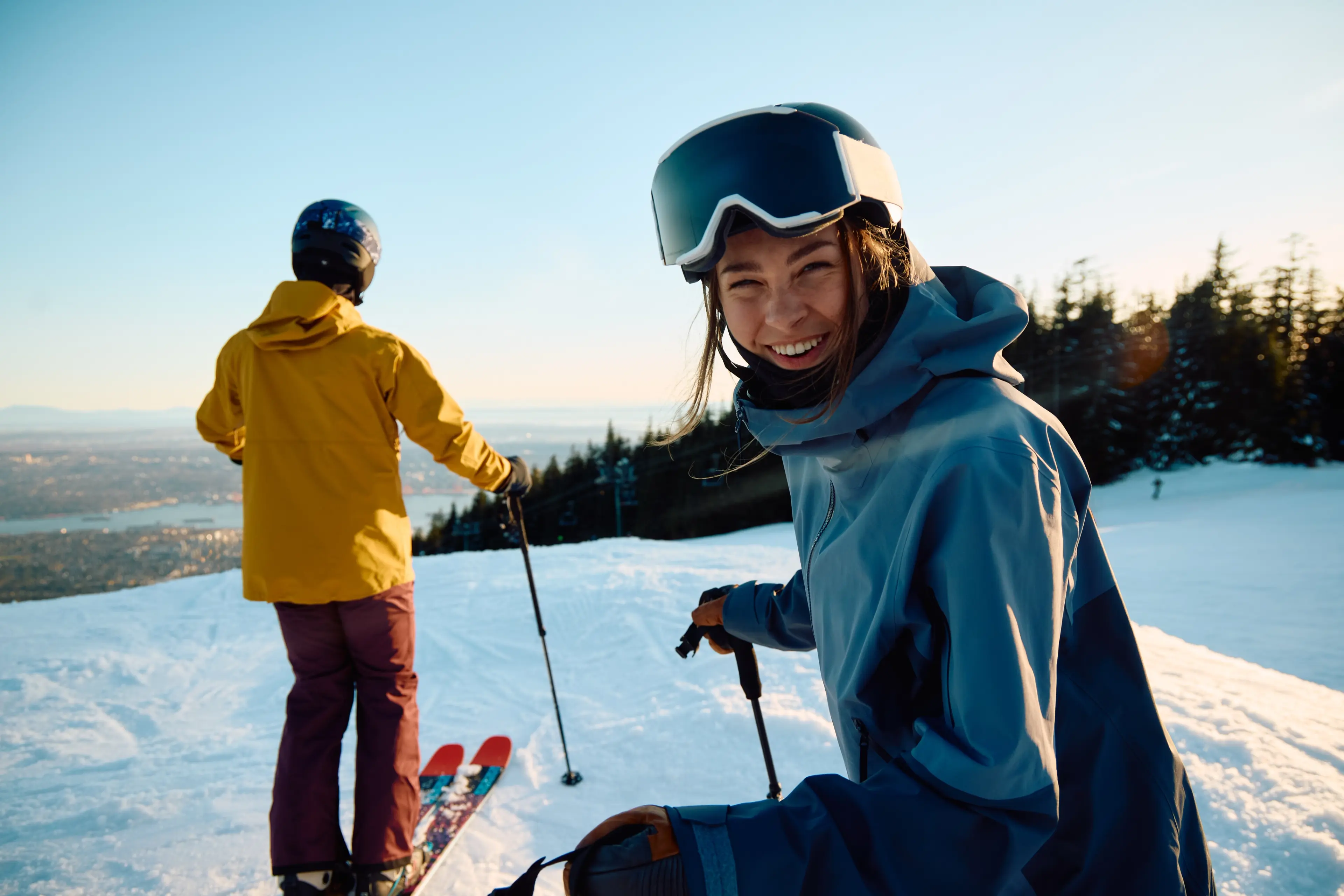 Two skiers on a snowy slope at sunset, one smiling at the camera wearing gear and goggles, the other facing away in a ski jacket.
