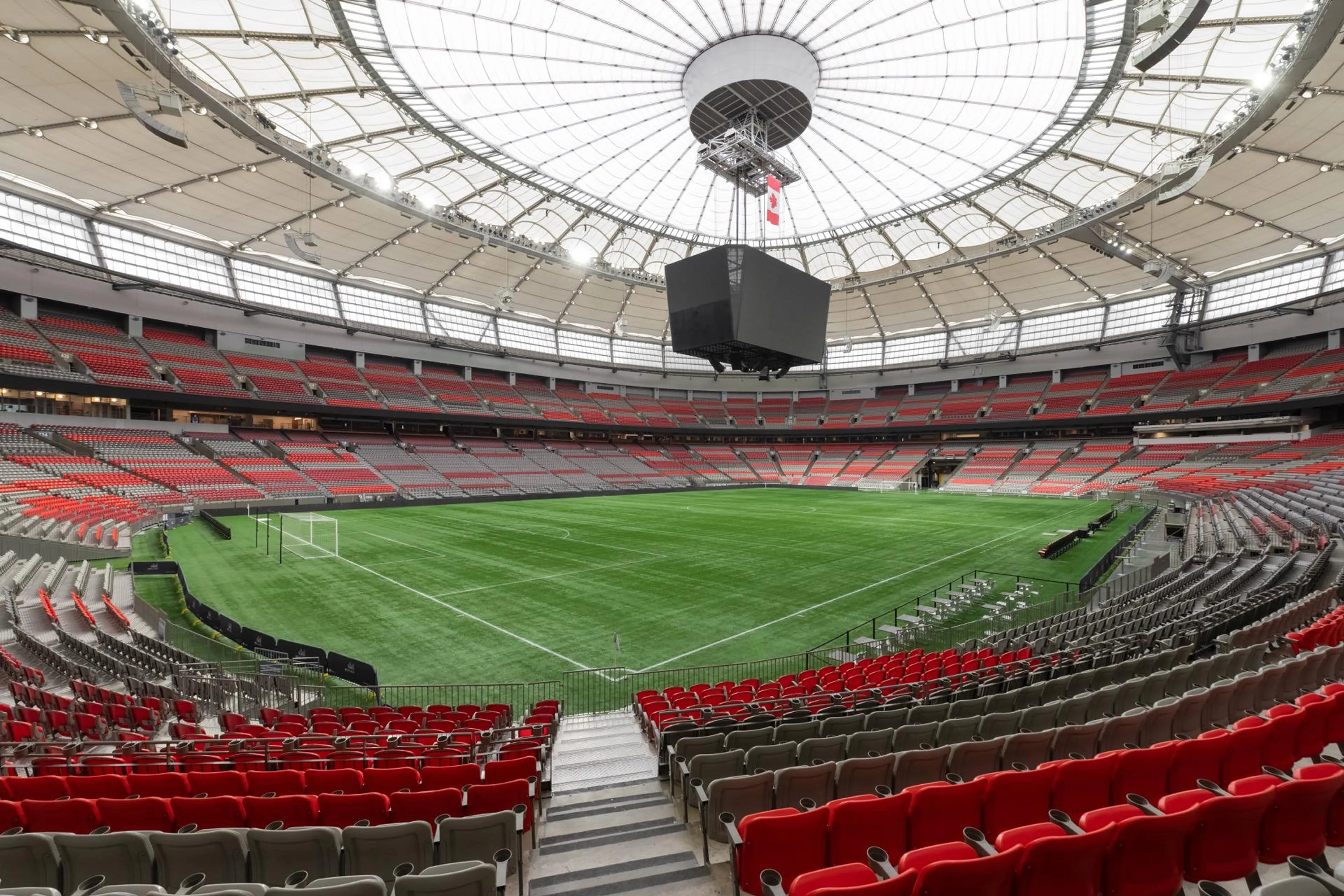 Empty modern stadium with red and gray seats, green soccer field, and large central scoreboard under a white roof.