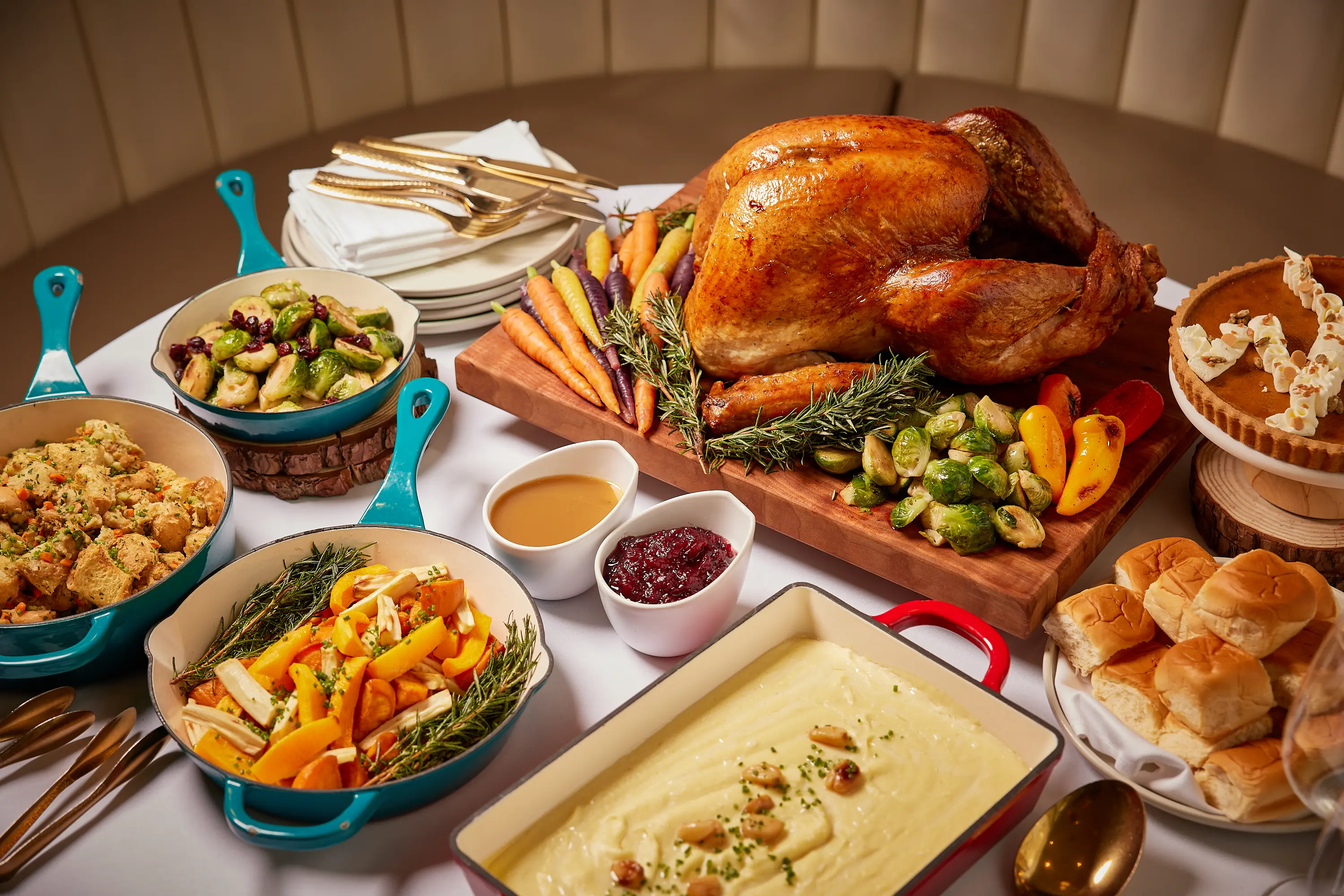 Roasted turkey on a wooden board with carrots, Brussels sprouts, and herbs surrounded by bowls of stuffing, roasted vegetables, mashed potatoes, gravy, cranberry sauce, dinner rolls, and a pumpkin pie.
