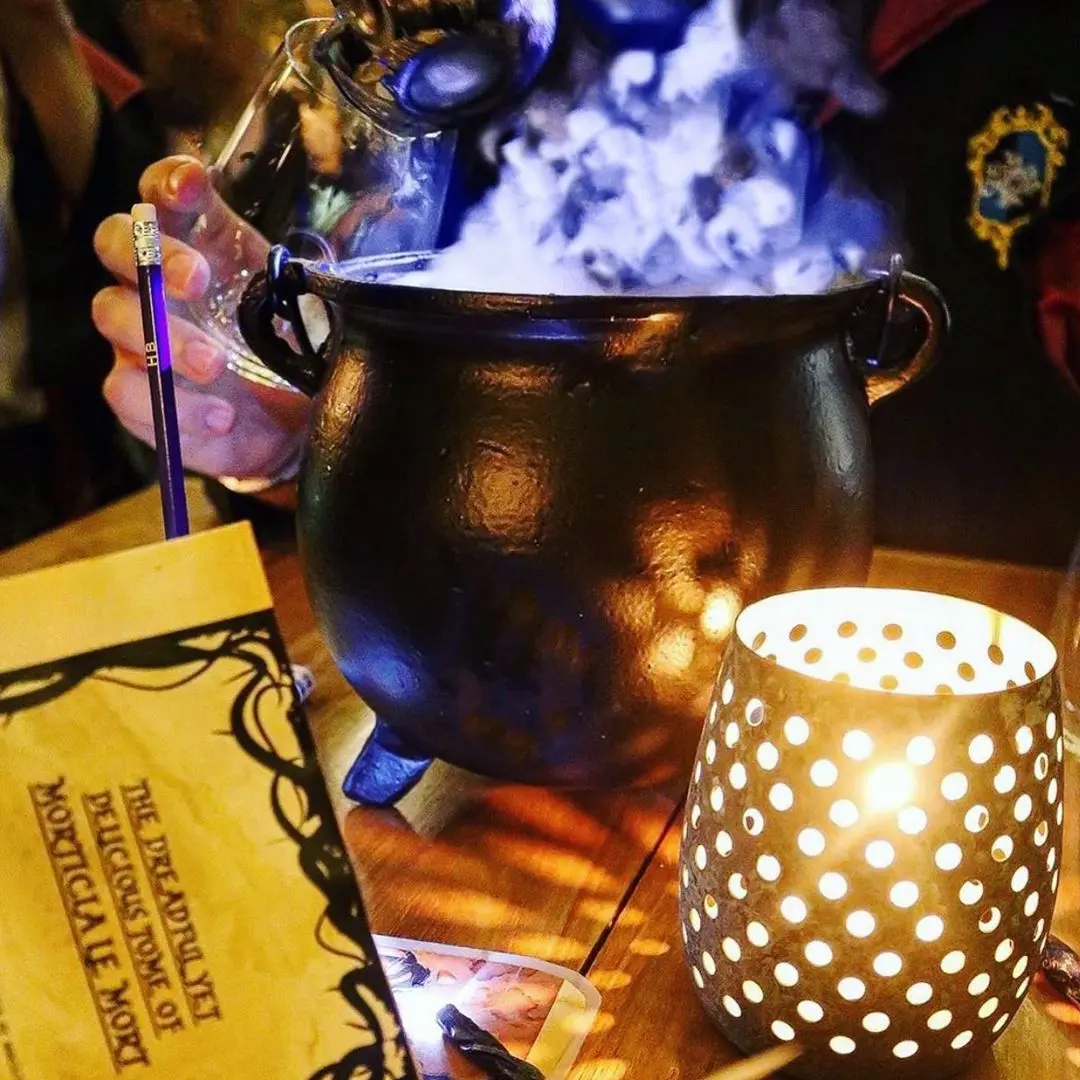 A hand pouring liquid into a smoking black cauldron on a wooden table with a lit candle holder and a menu titled 'The Deadfully Delicious Tome of Morticale Moet'.