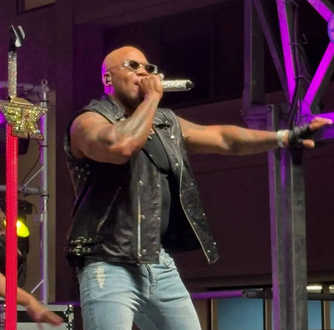Bald man wearing sunglasses, a black sleeveless leather jacket, and ripped jeans singing into a microphone on stage.