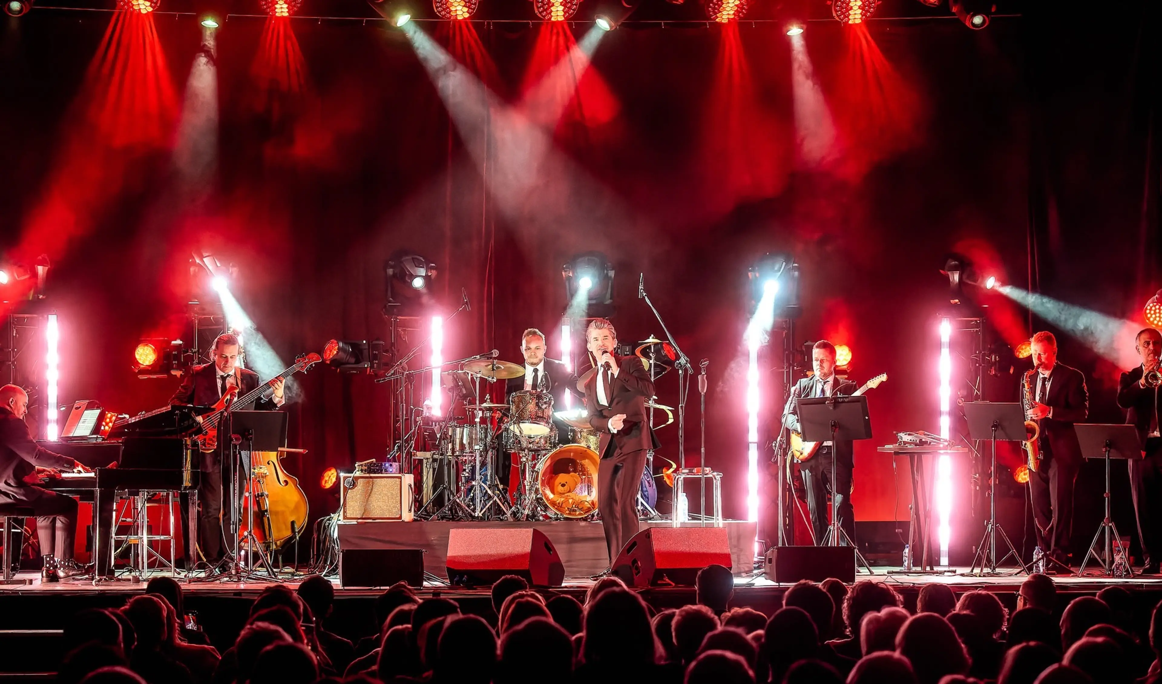 A seven-person band performing on stage with red lighting, including a singer, drummer, pianist, guitarist, bassist, saxophonist, and trumpeter.