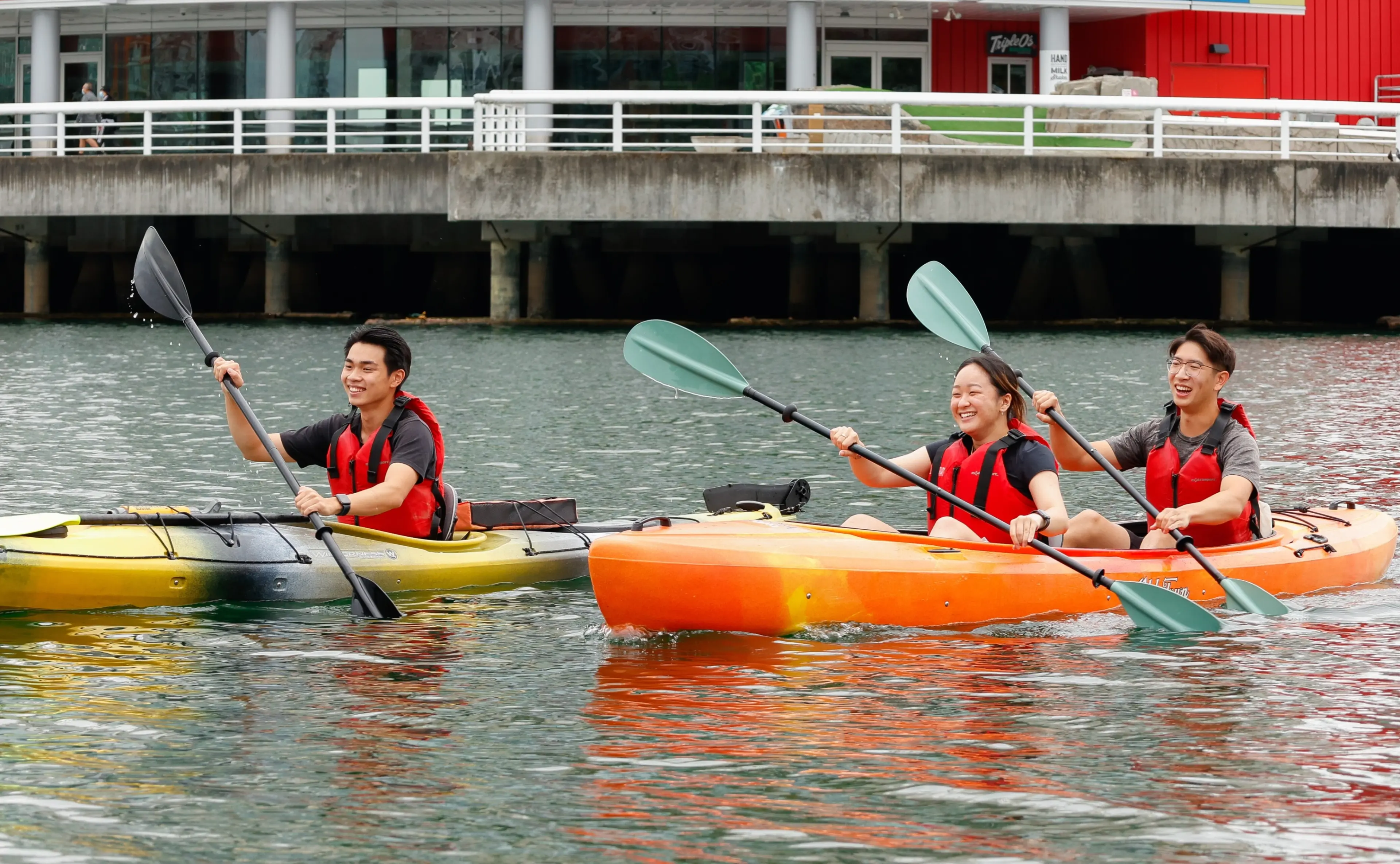 Three people wearing red life jackets kayaking on calm water near a dock with buildings in the background.