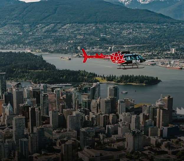 Red and black helicopter flying over a city with tall buildings, water, and forested hills in the background.