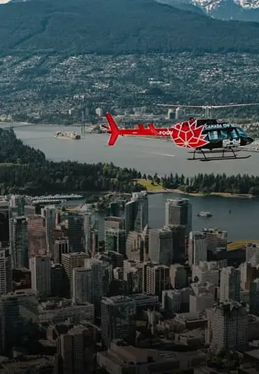Red and black helicopter flying over a city with tall buildings, water, and forested hills in the background.