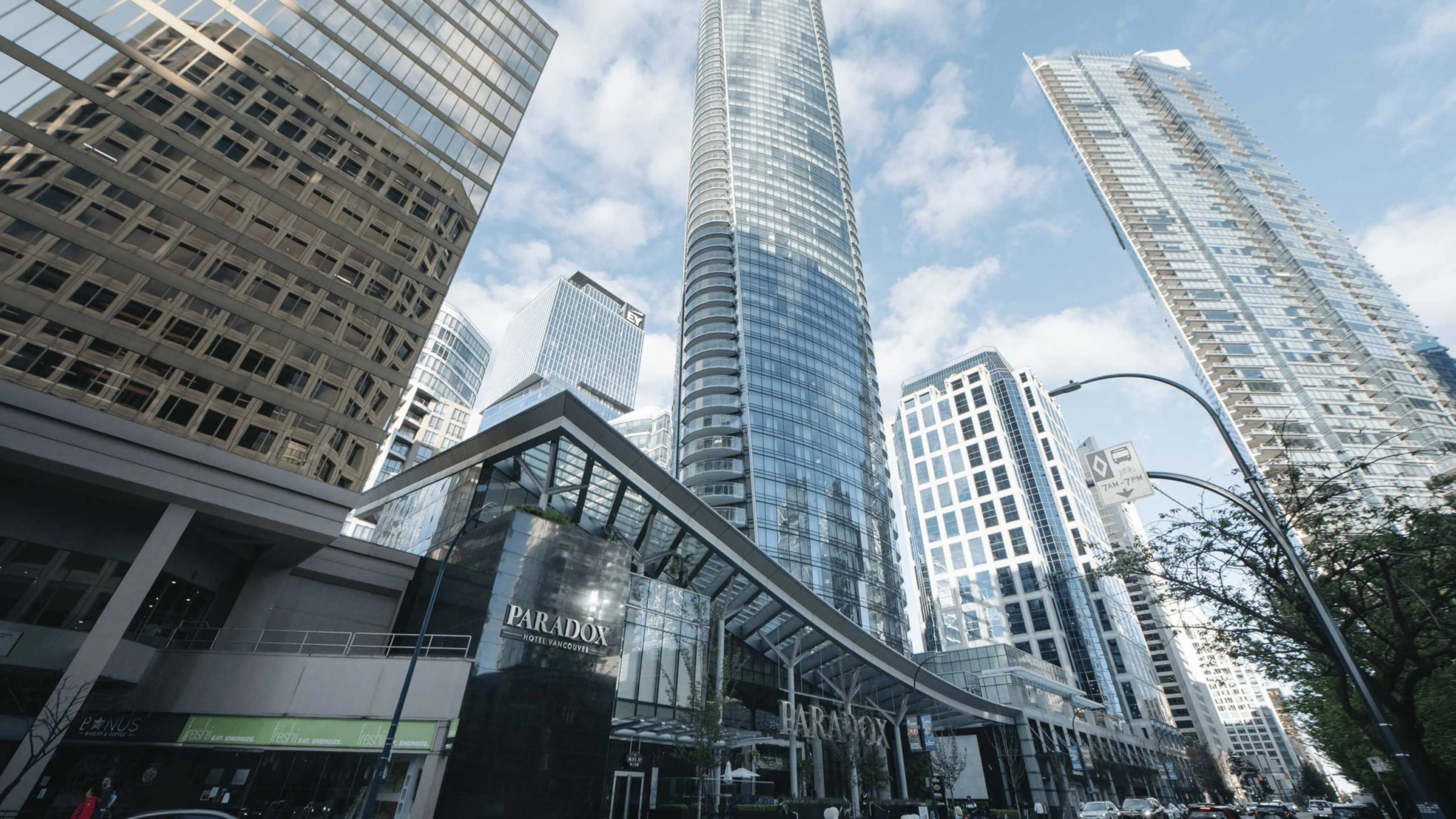 Tall modern glass skyscrapers surrounding the Paradox Hotel Vancouver on a partly cloudy day.