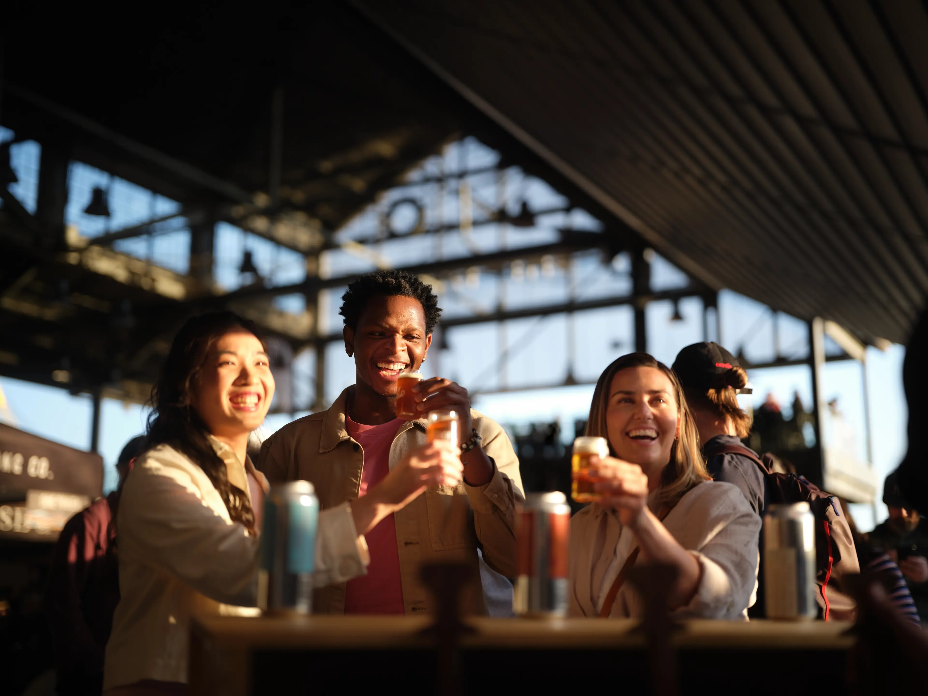 Three people smiling and holding up glasses of beer in a bright indoor setting with metal beams overhead.