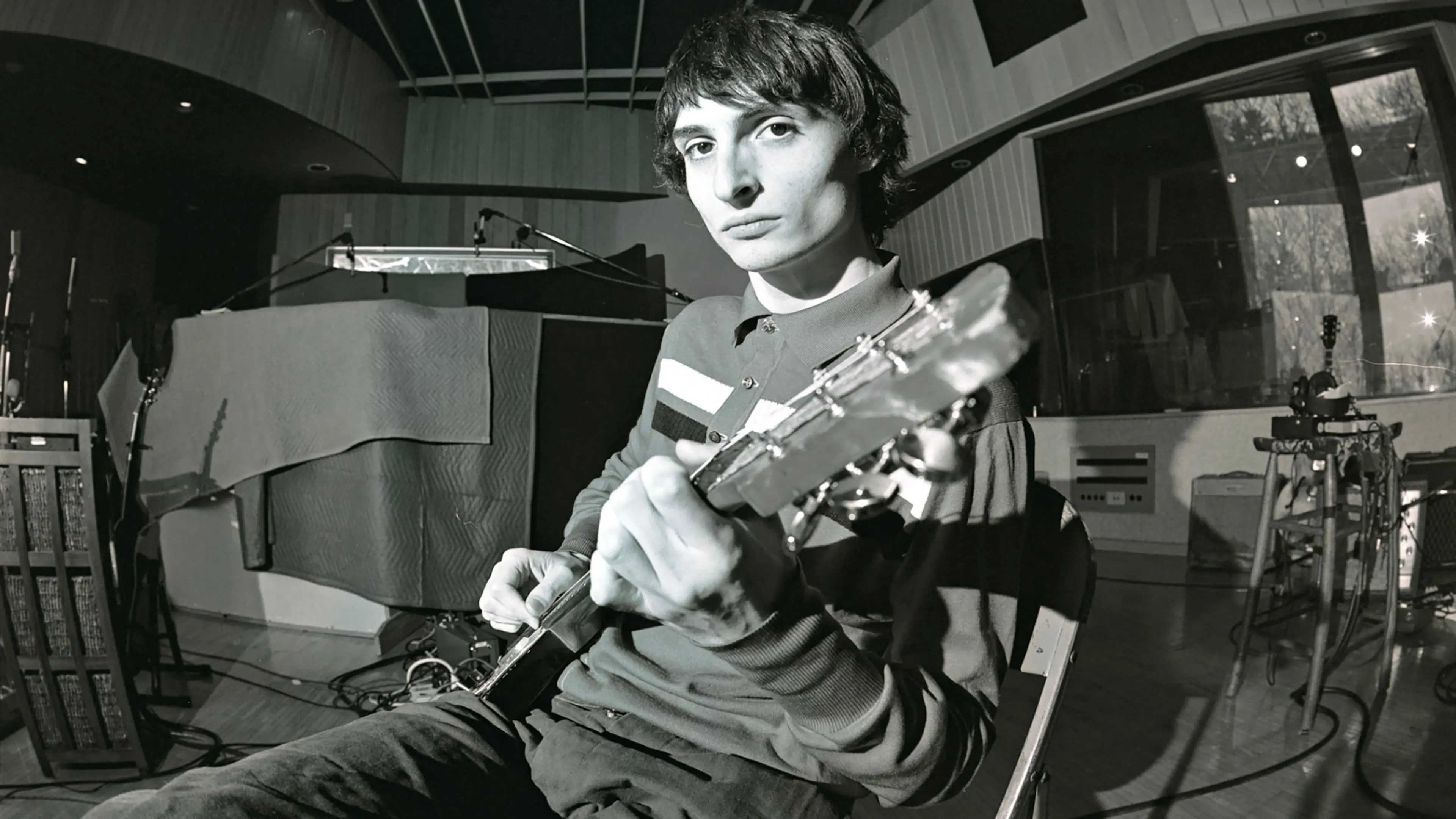 Person sitting in a recording studio playing an electric guitar, looking toward the camera.