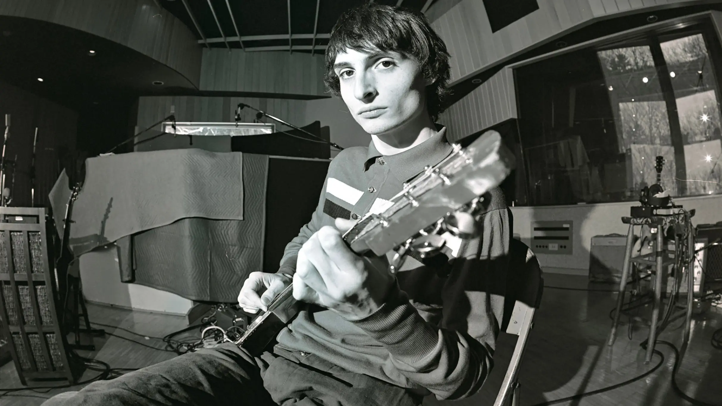 Person sitting in a recording studio playing an electric guitar, looking toward the camera.