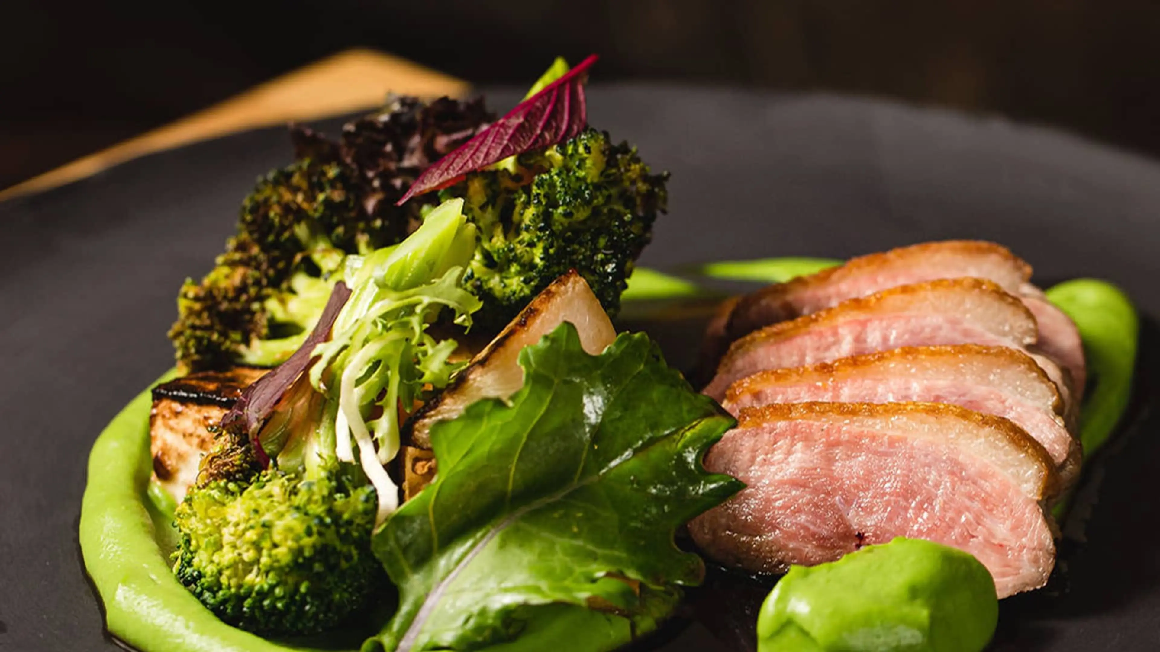 Sliced cooked meat served with grilled broccoli and leafy greens on a black plate with green sauce.