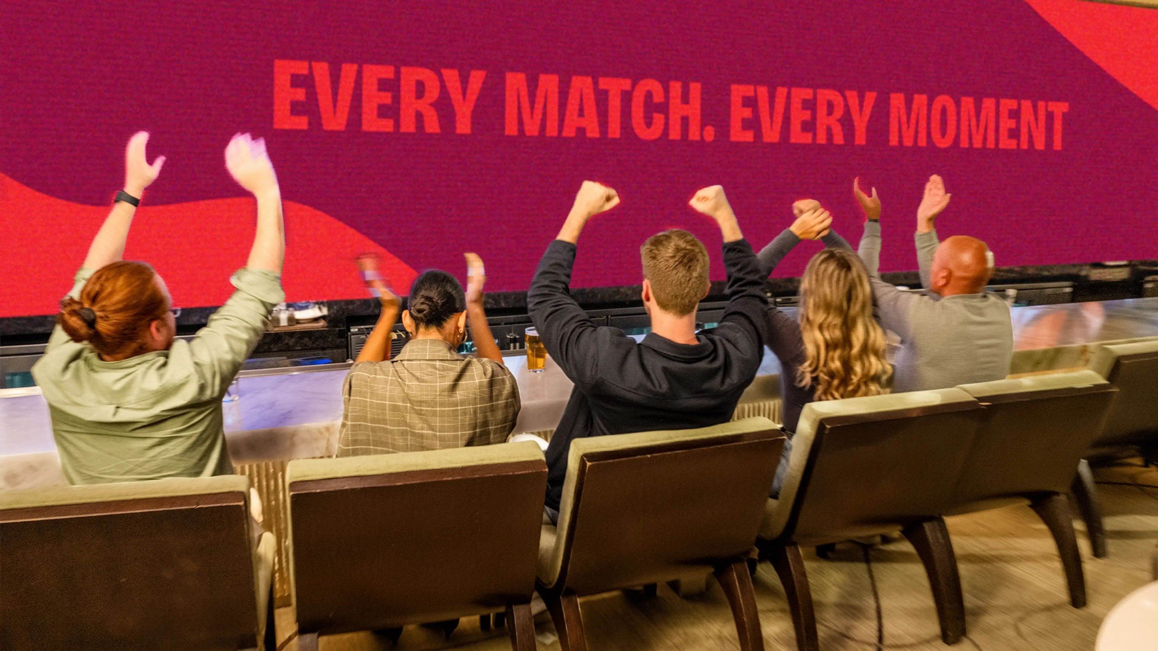 Five people sitting in chairs facing a large screen with text Every Match. Every Moment, cheering with raised hands.