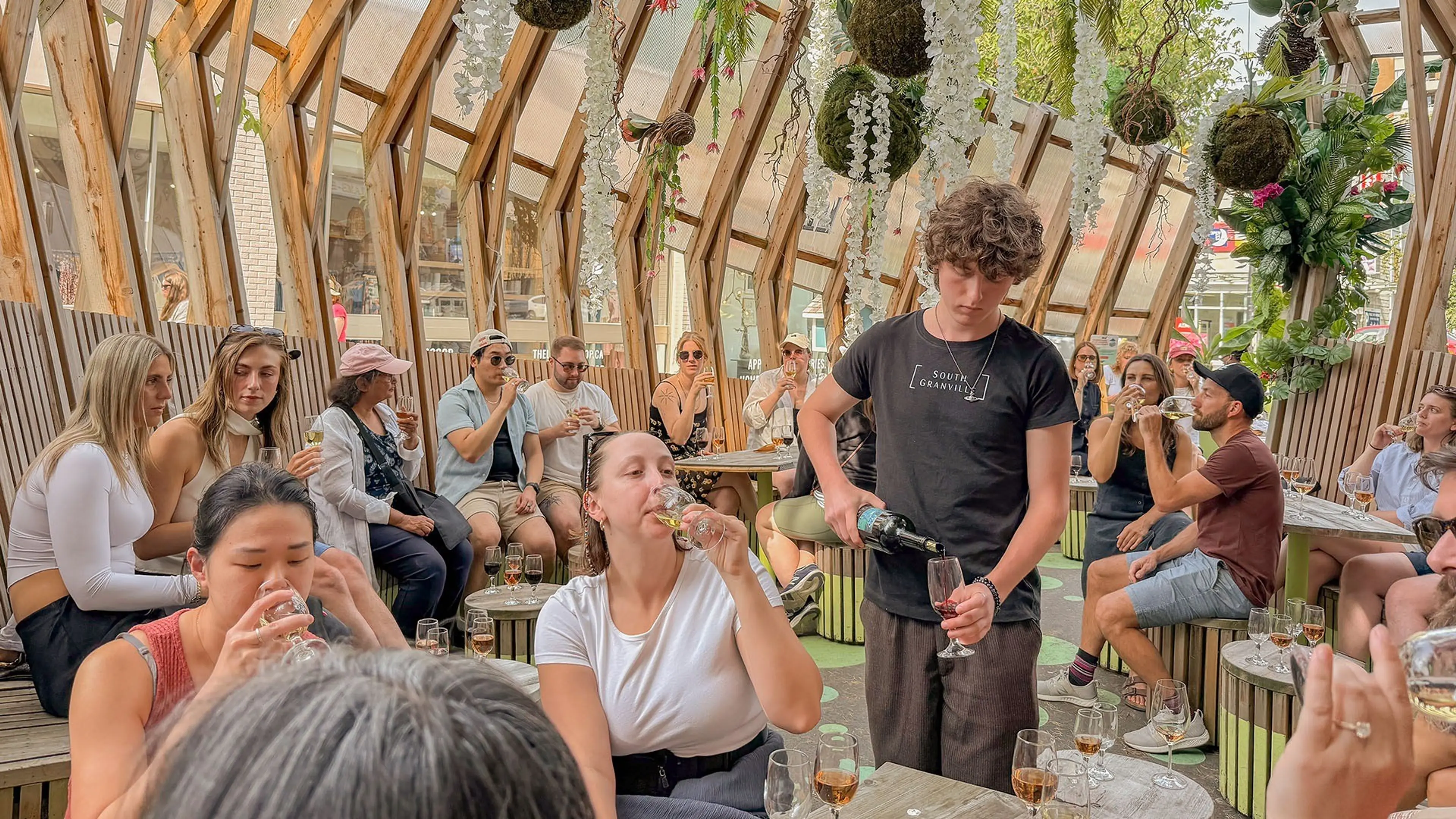A person pouring wine for a seated person drinking from a glass in a wooden indoor garden café with hanging plants and multiple people seated and drinking.