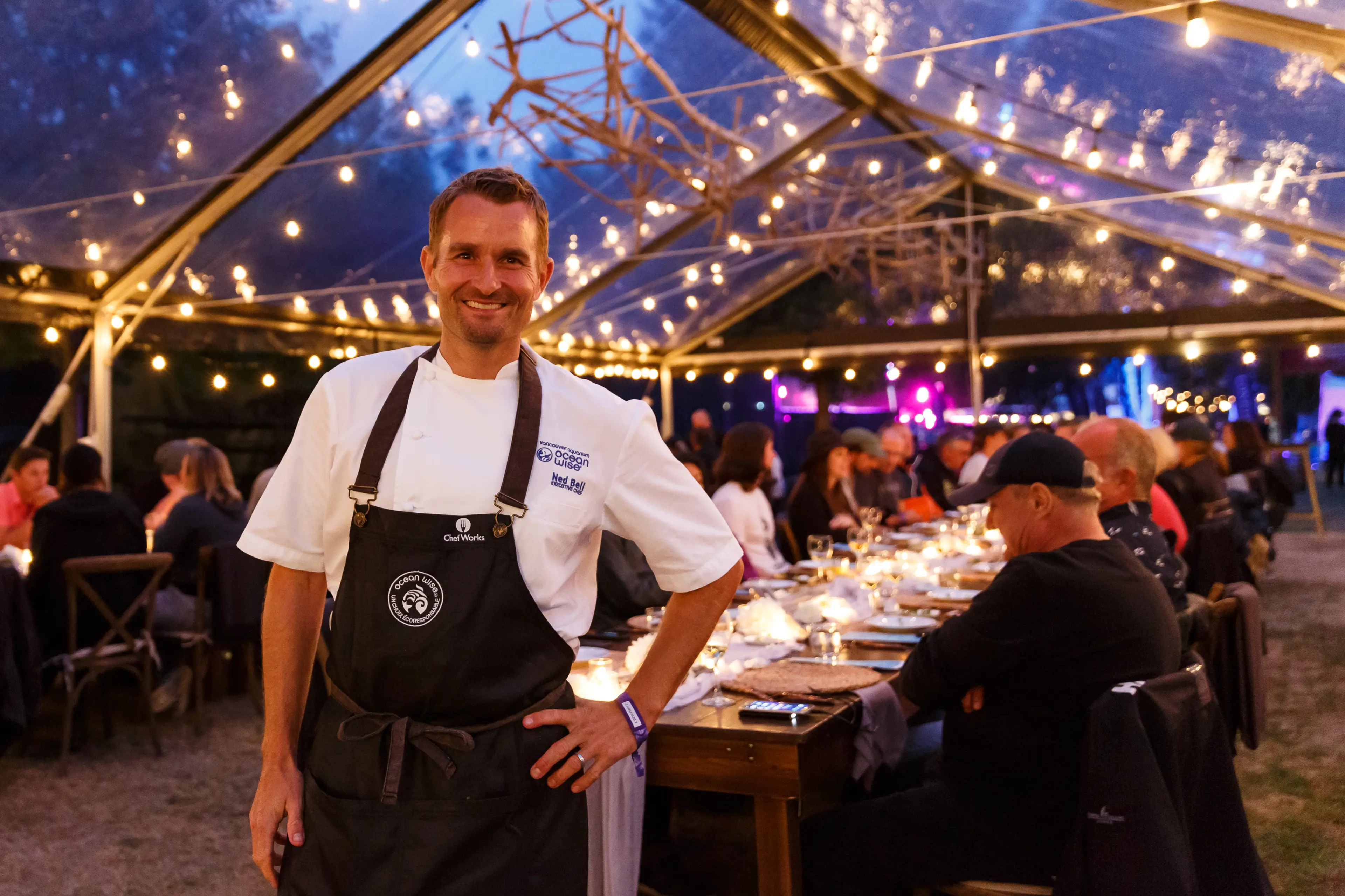 Chef Ned Bell with an Ocean Wise apron and chef's jacket posing in from of a SKOOKUM Music Festival Long Table Dinner
