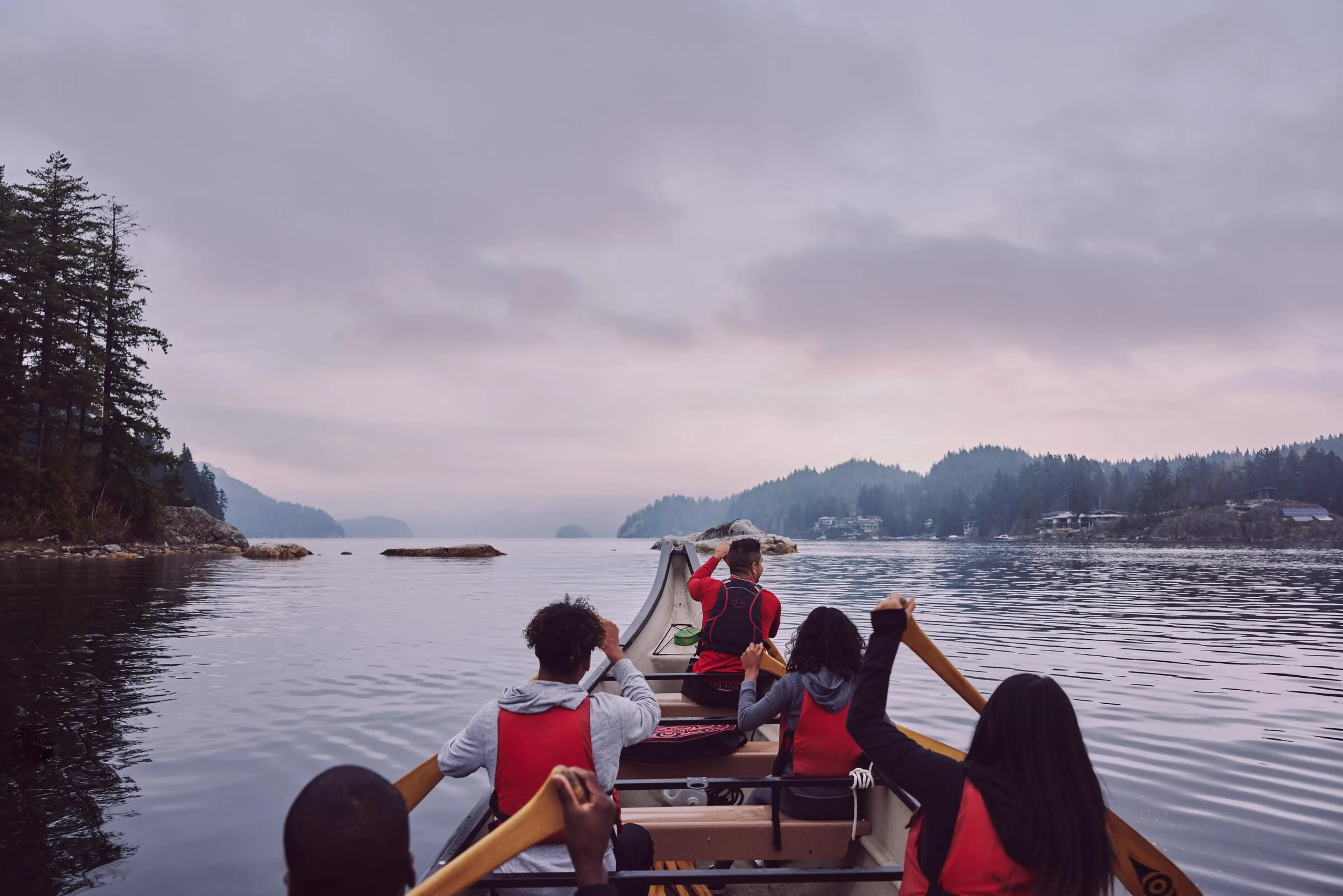 Five people wearing life jackets paddling a canoe on calm water surrounded by forested hills under a cloudy sky.