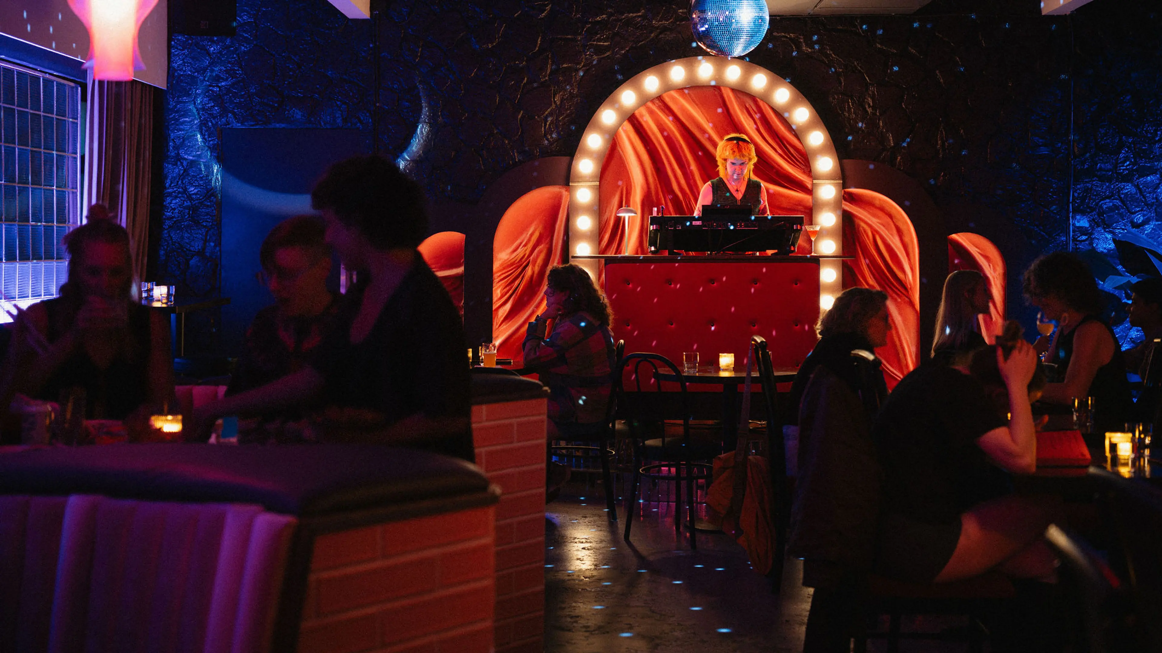 DJ playing music on a red-lit stage with people sitting and socializing at tables in a dimly lit bar or club.
