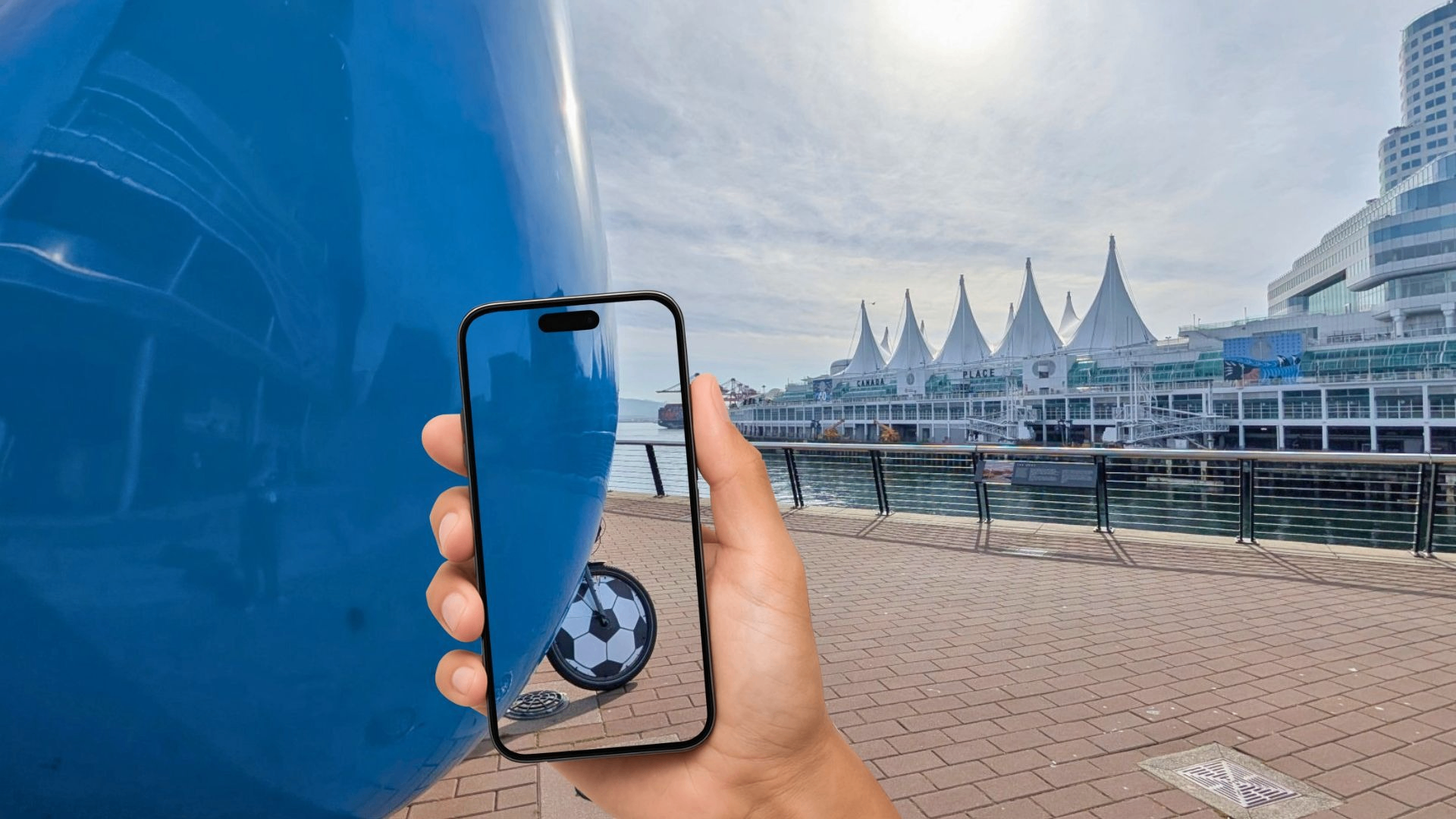 Hand holding a smartphone showing a soccer ball behind a large blue object on a waterfront promenade with white tent-like structures in the background.