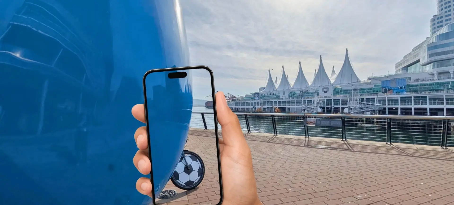 Hand holding a smartphone showing a soccer ball behind a large blue object on a waterfront promenade with white tent-like structures in the background.