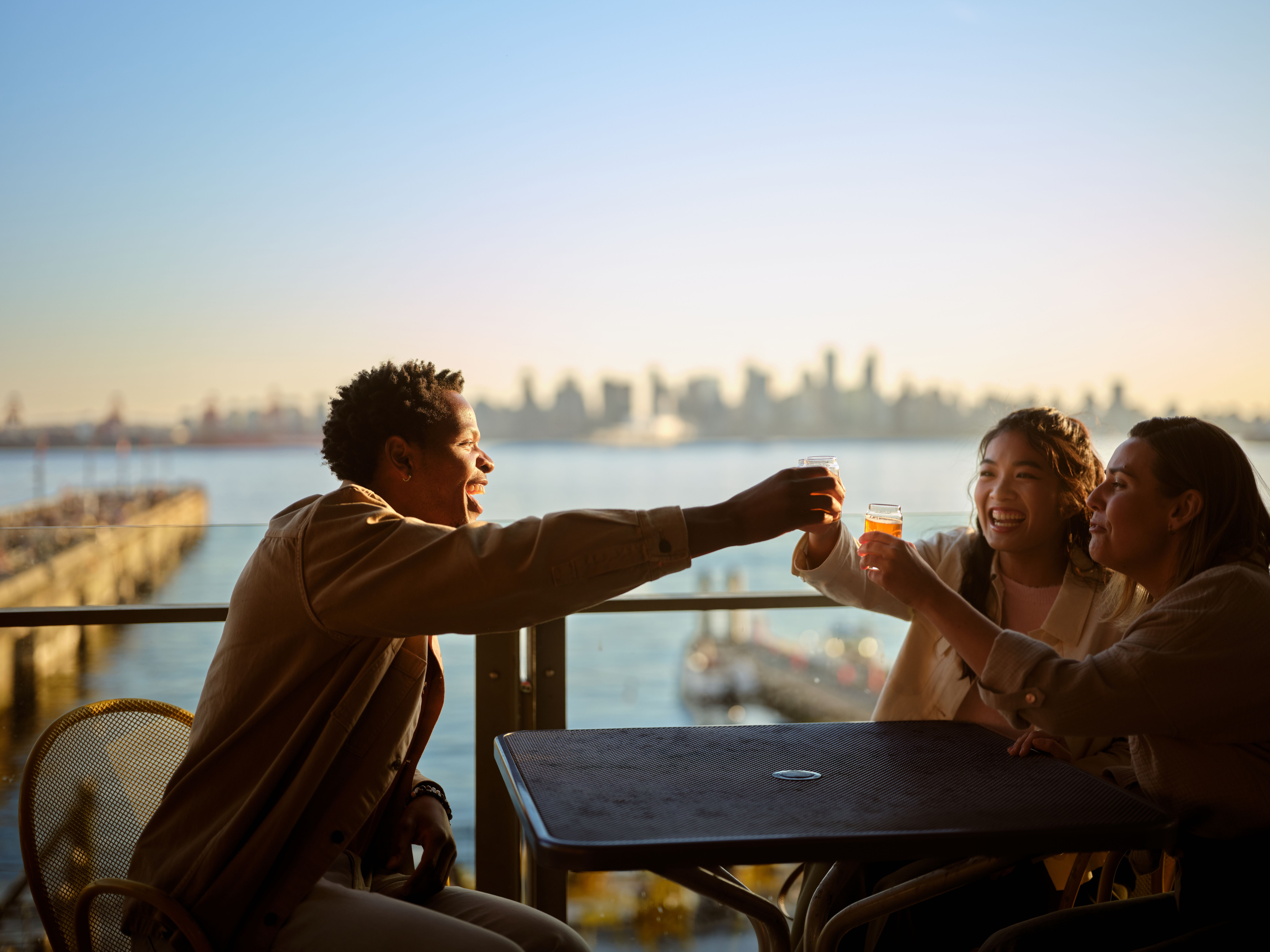 Three friends on a patio cheering with drinks, with a view of the ocean and downtown in the distance.
