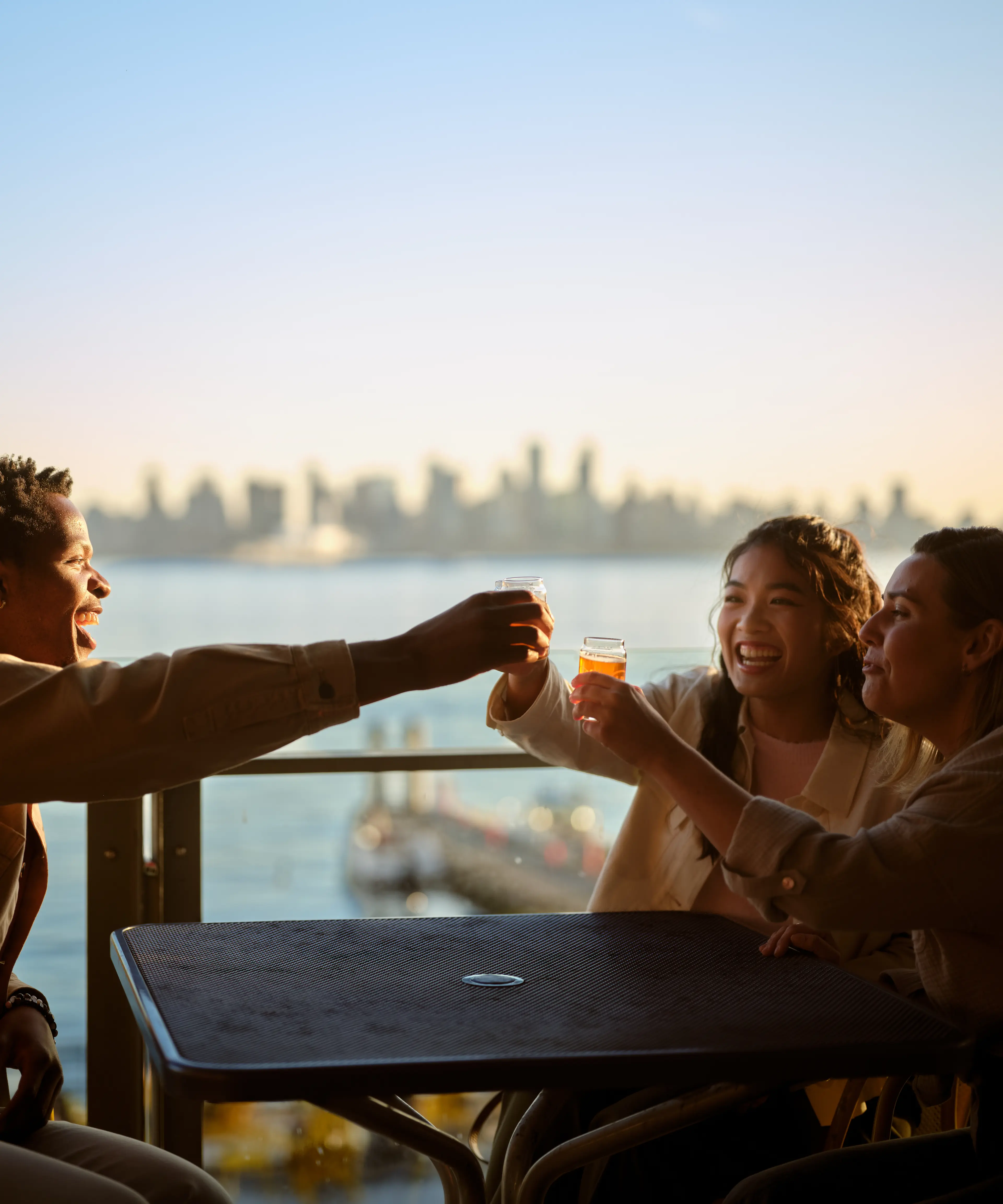 Three friends on a patio cheering with drinks, with a view of the ocean and downtown in the distance.