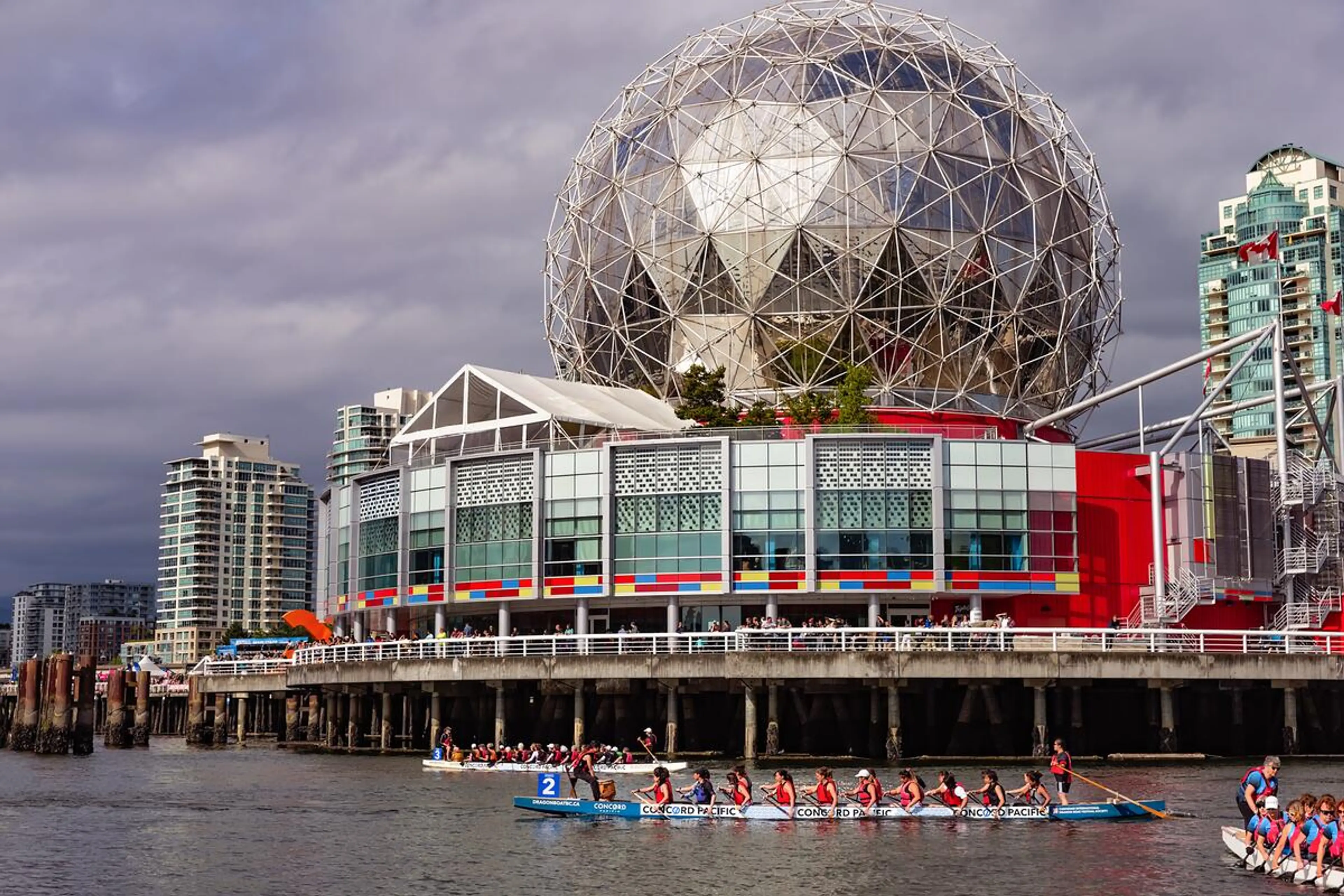 Two dragon boats with paddlers on water near a modern building with a large geodesic dome and city skyline in the background.