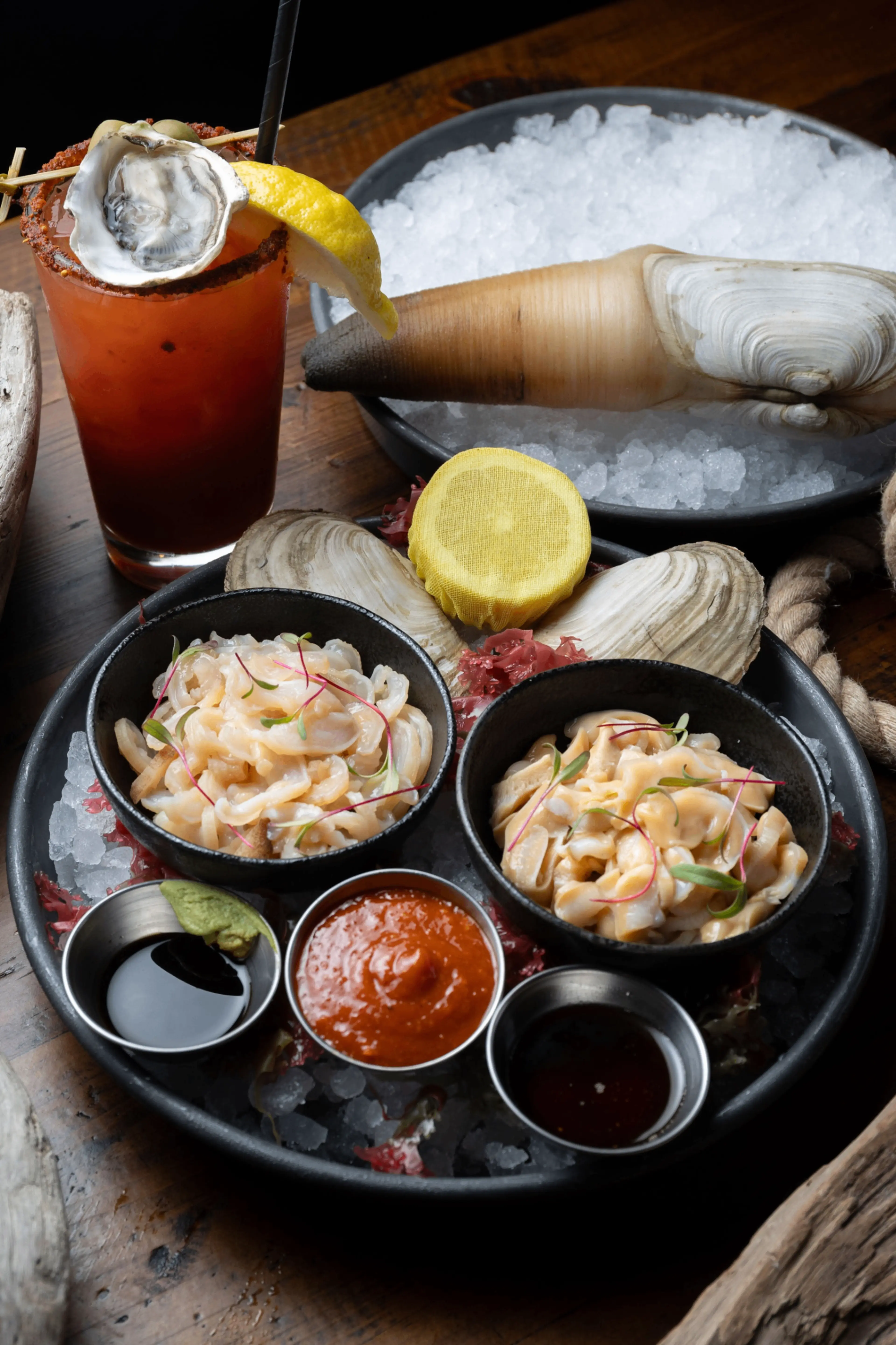 Seafood platter with two bowls of shellfish salad, three dipping sauces, lemon slice, and a cocktail garnished with an oyster and lemon wedge.