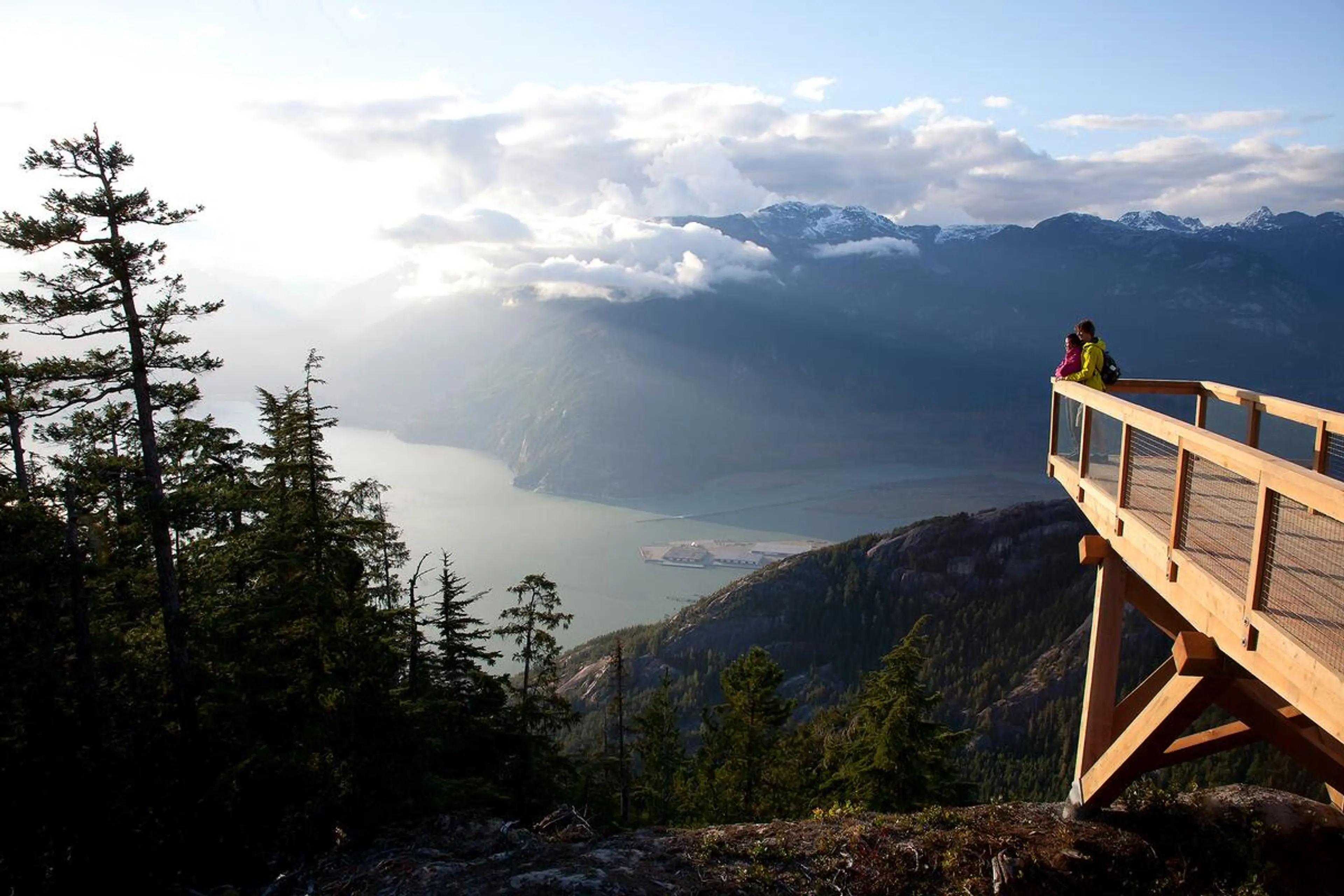 Two people standing on a wooden viewing platform overlooking a forested mountain landscape and a body of water with clouds and sunlight.