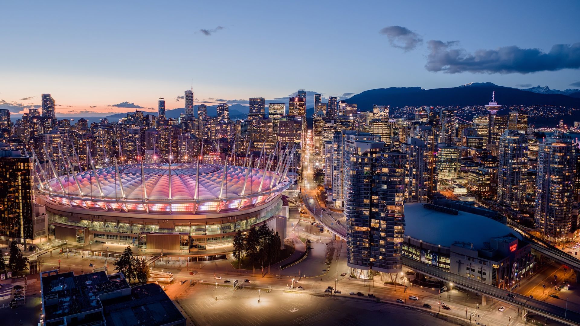 Aerial view of a city skyline at dusk with a large stadium lit in pink and white lights in the foreground and tall buildings surrounding it.