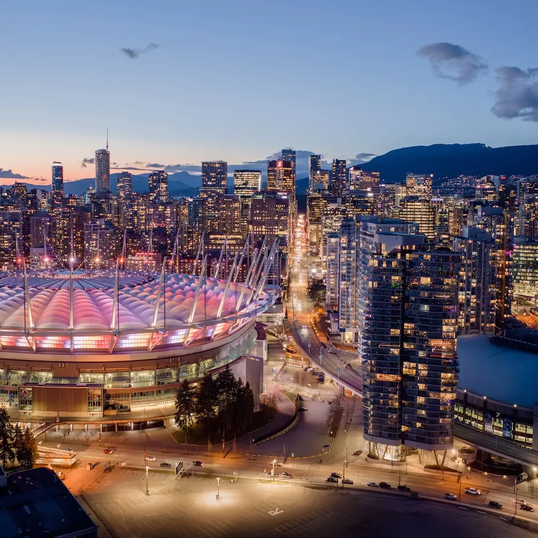 Aerial view of a city skyline at dusk with a large stadium lit in pink and white lights in the foreground and tall buildings surrounding it.