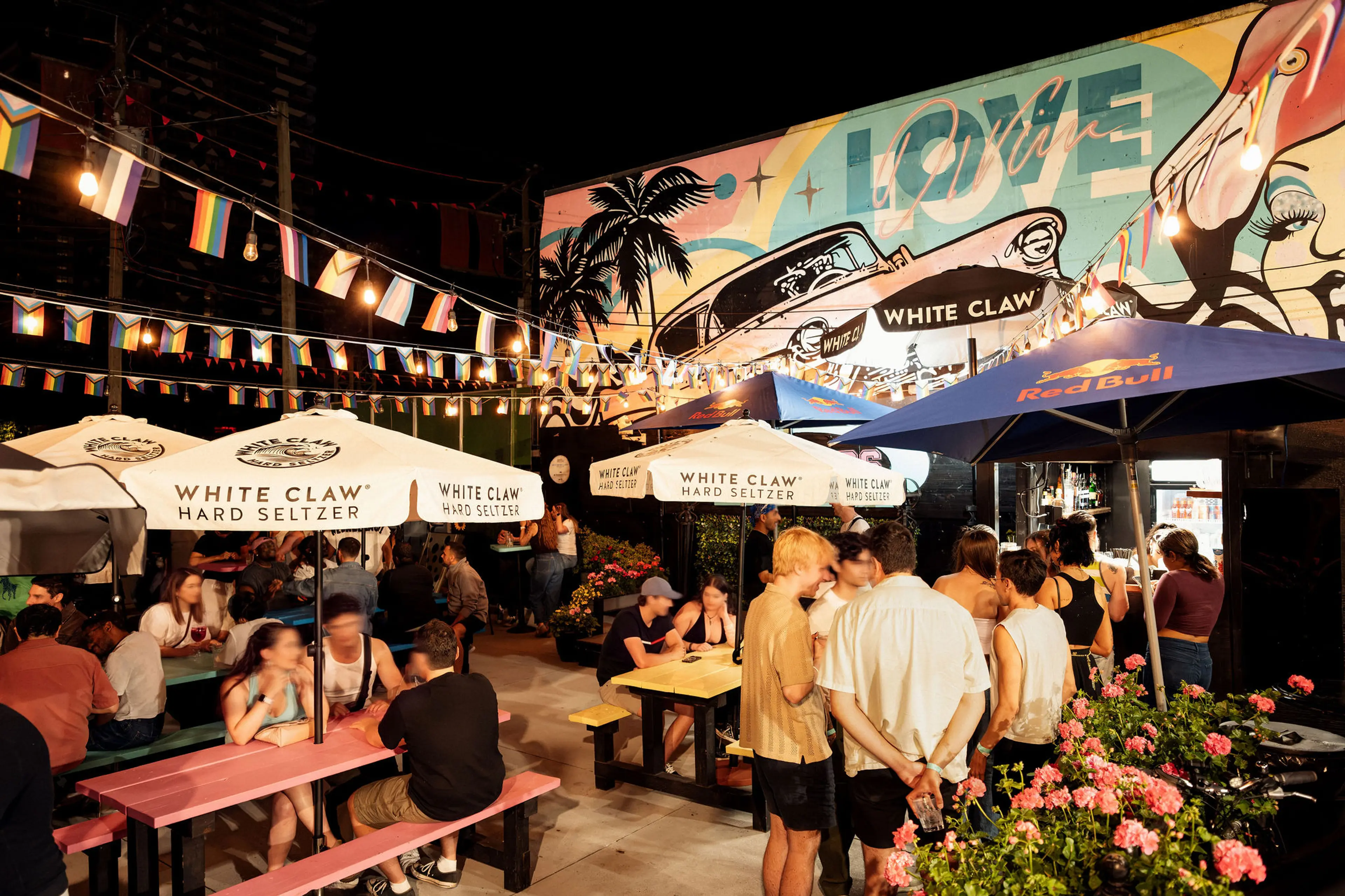 Outdoor night scene with people socializing under White Claw and Red Bull umbrellas, colorful flags, and a mural with the word LOVE.