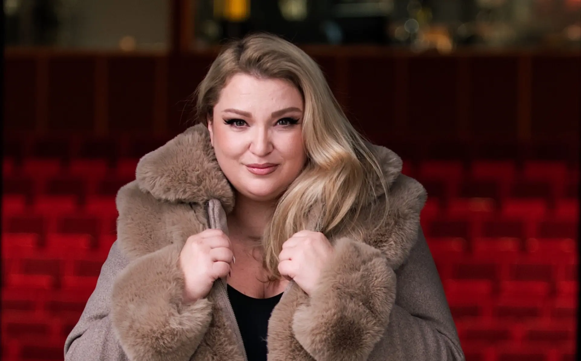 Person with long blonde hair wearing a large brown fur coat, holding the collar, with red theater seats in the background.