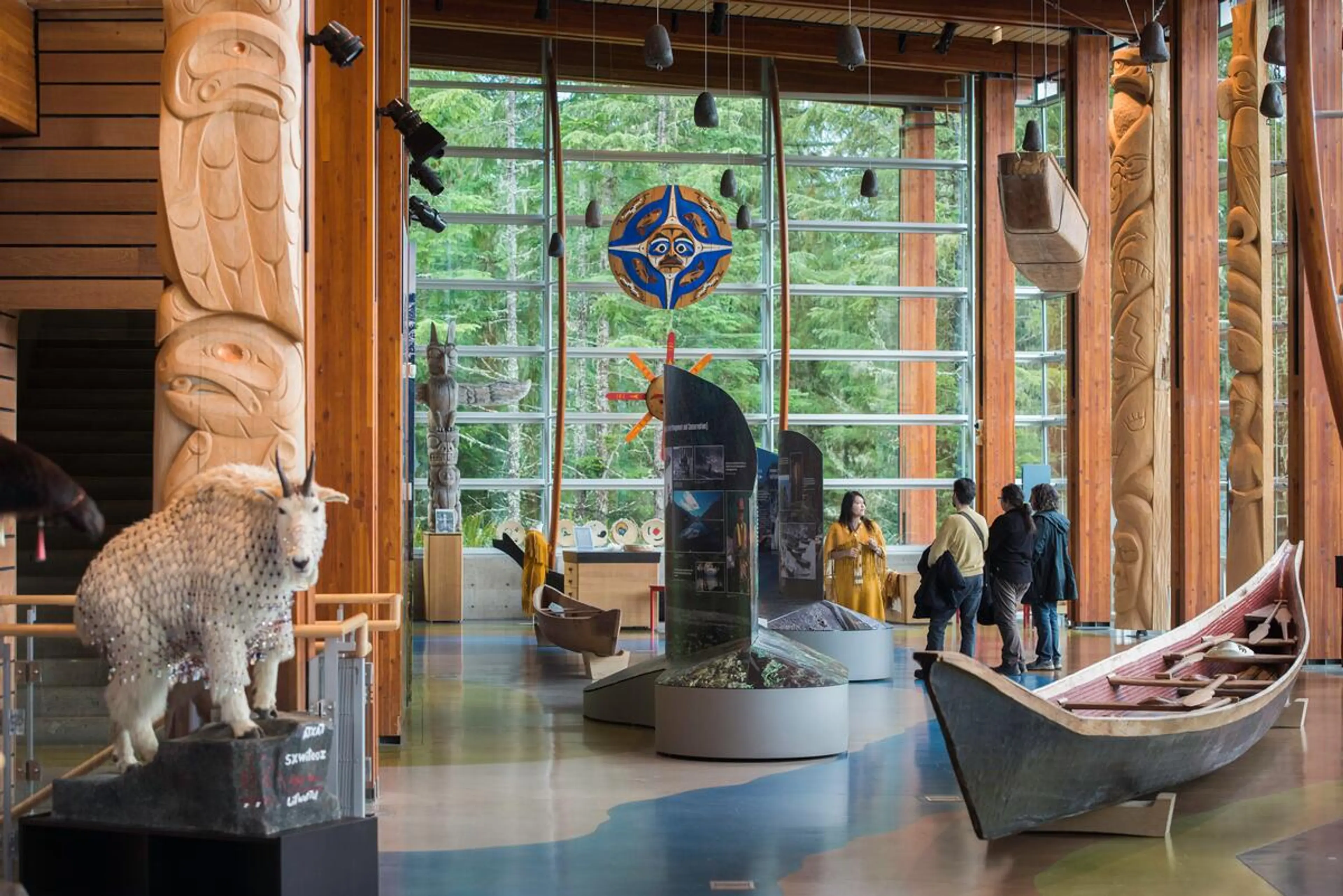 Museum interior with wooden totem poles, a carved wooden canoe, a taxidermy mountain goat, and four people near exhibit displays.