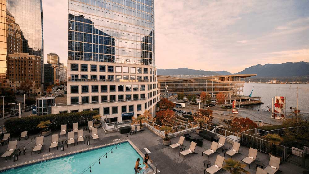 Outdoor pool with lounge chairs on a rooftop terrace overlooking city buildings, waterfront, and mountains at sunset.
