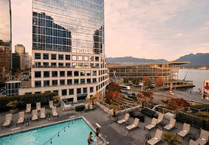 Outdoor pool with lounge chairs on a rooftop terrace overlooking city buildings, waterfront, and mountains at sunset.