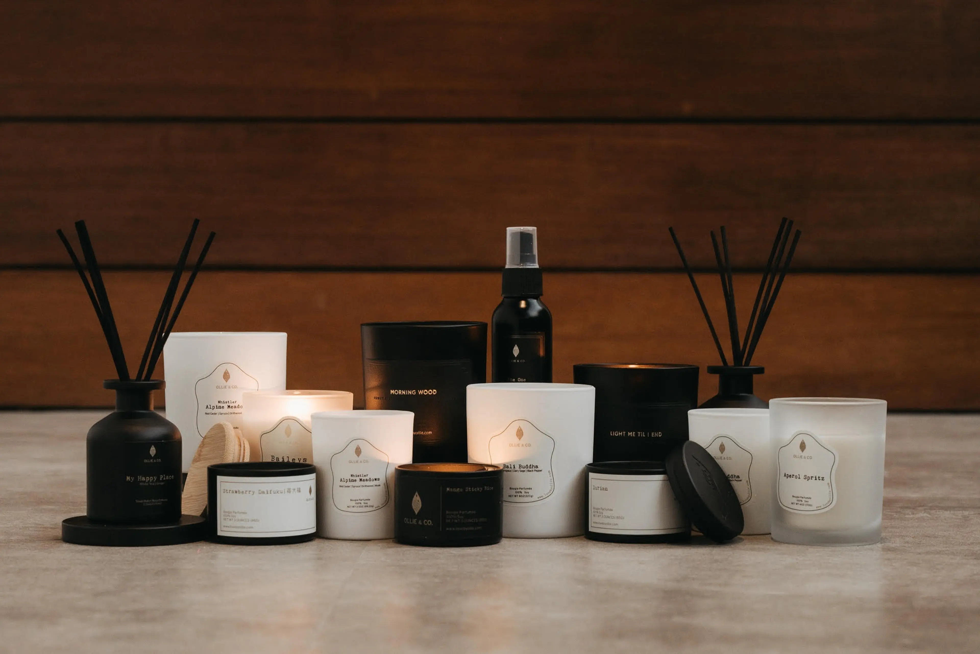 A collection of black and white scented candles and reed diffusers arranged on a marble surface with a wooden background.