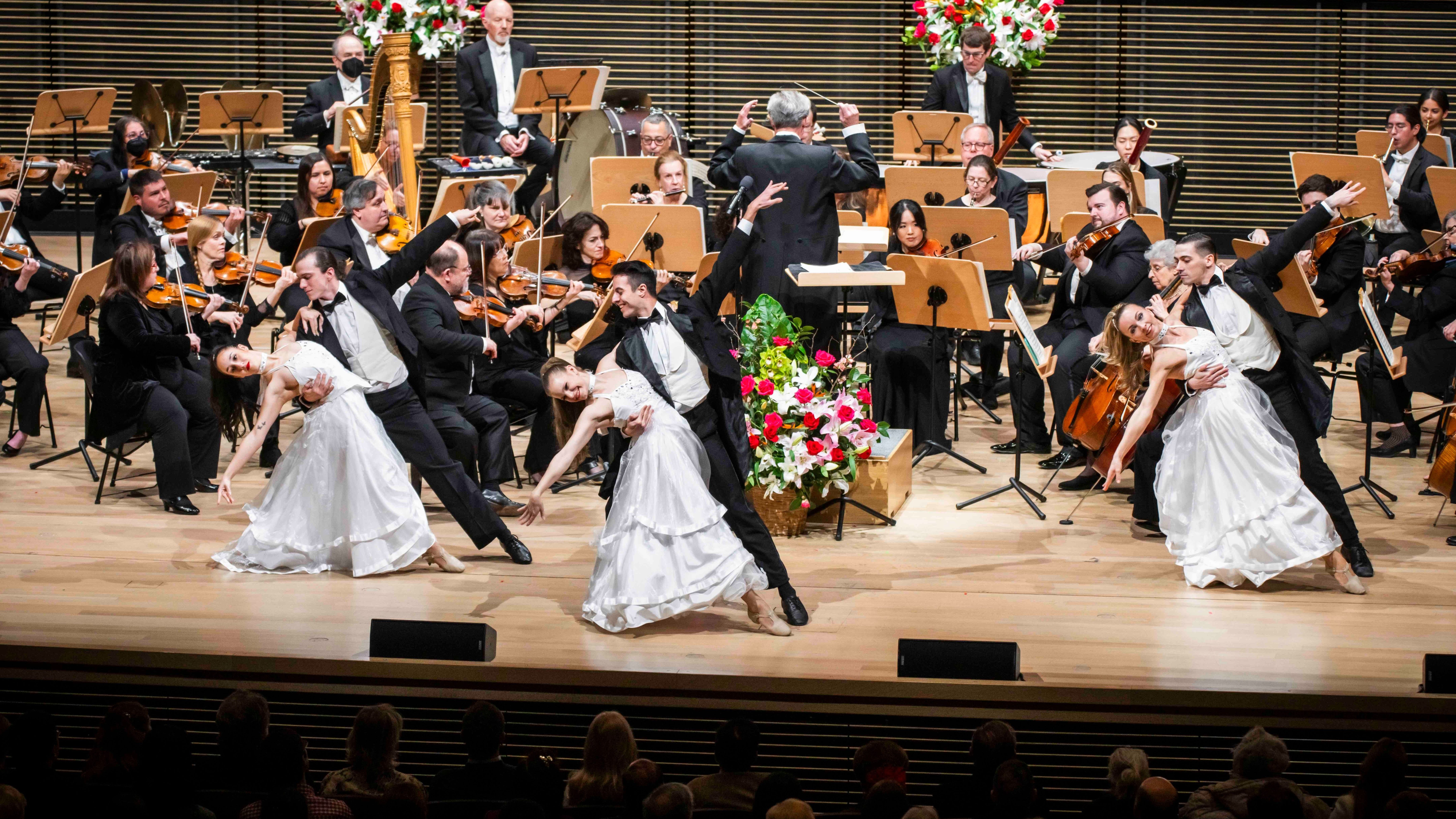 Three pairs of dancers in white dresses and black suits performing in front of an orchestra on a wooden stage.