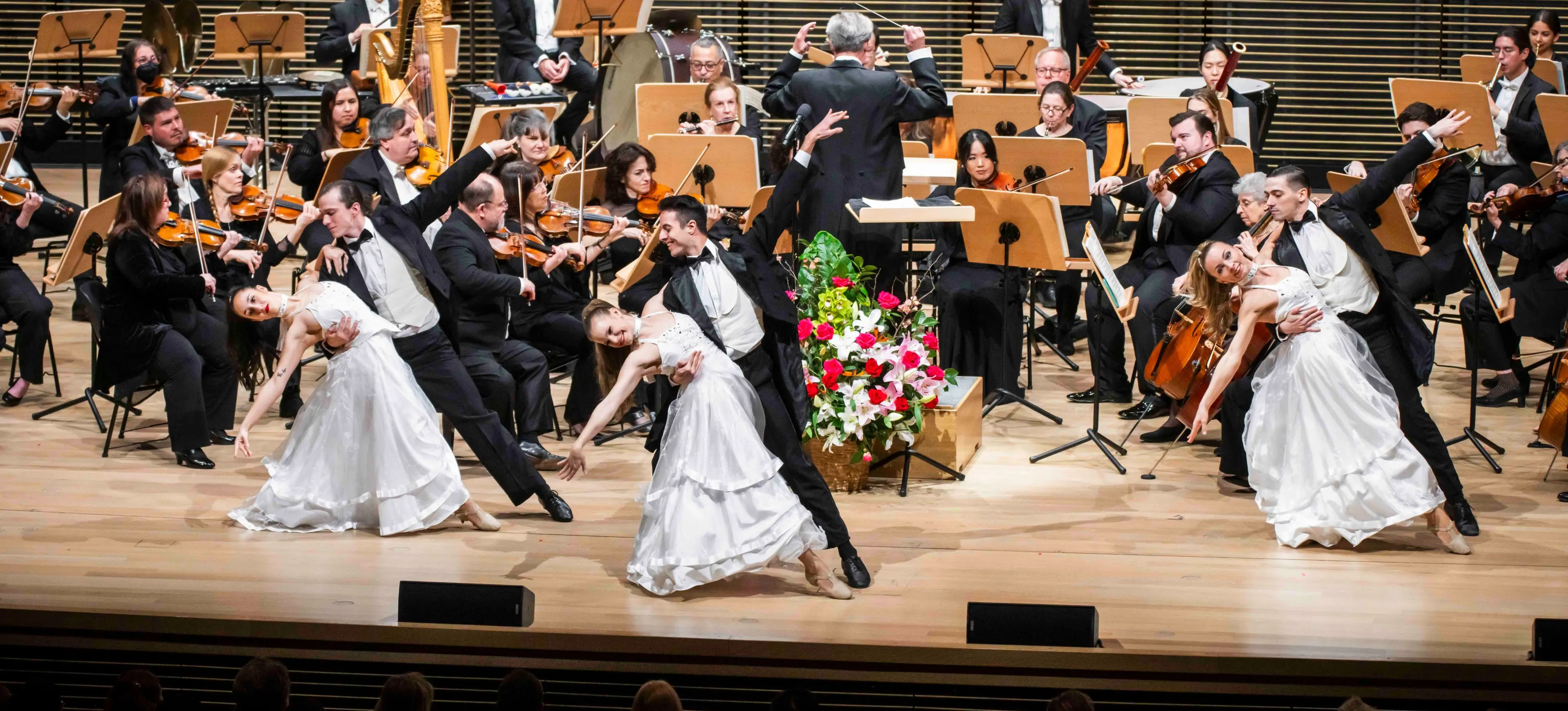 Three pairs of dancers in white dresses and black suits performing in front of an orchestra on a wooden stage.