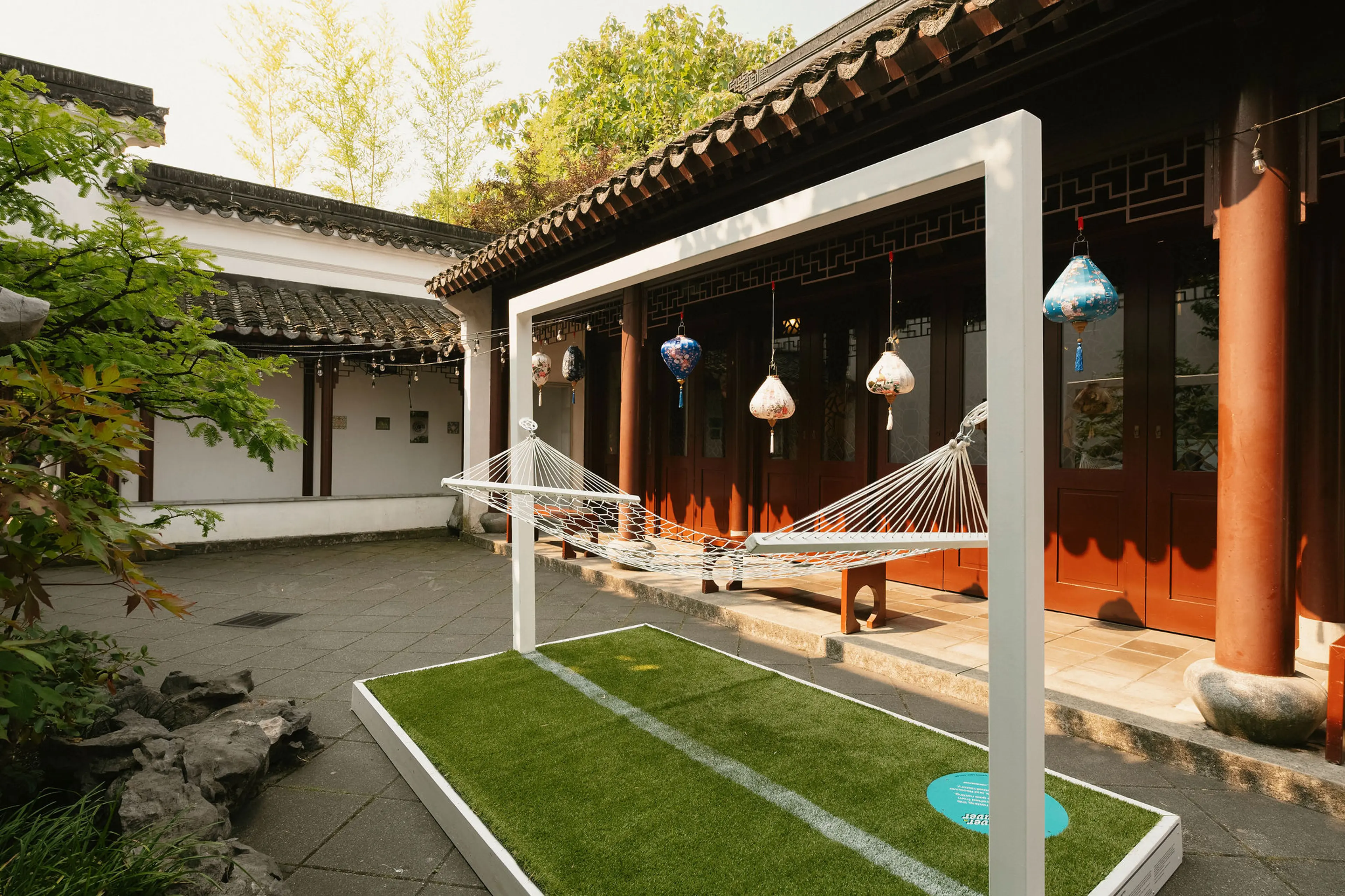 White hammock hanging on a white frame over a green artificial grass platform in a courtyard with traditional architecture and hanging lanterns.