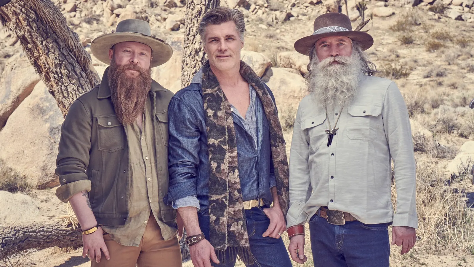 Three people standing outdoors in a rocky desert area, two wearing hats and long beards, one wearing a scarf and denim shirt.