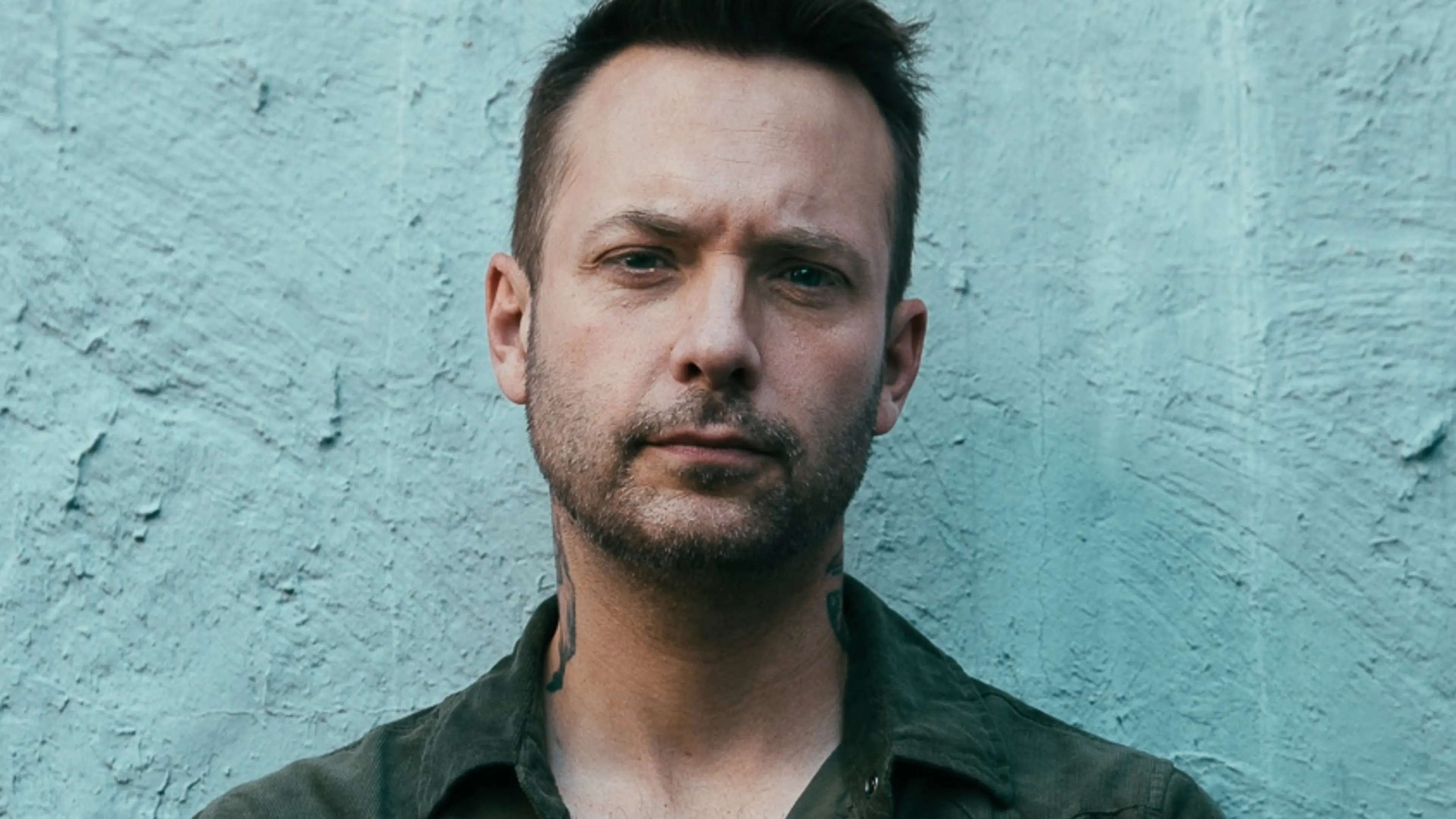 Person with short dark hair and light facial hair wearing a dark green shirt, standing against a textured blue wall.