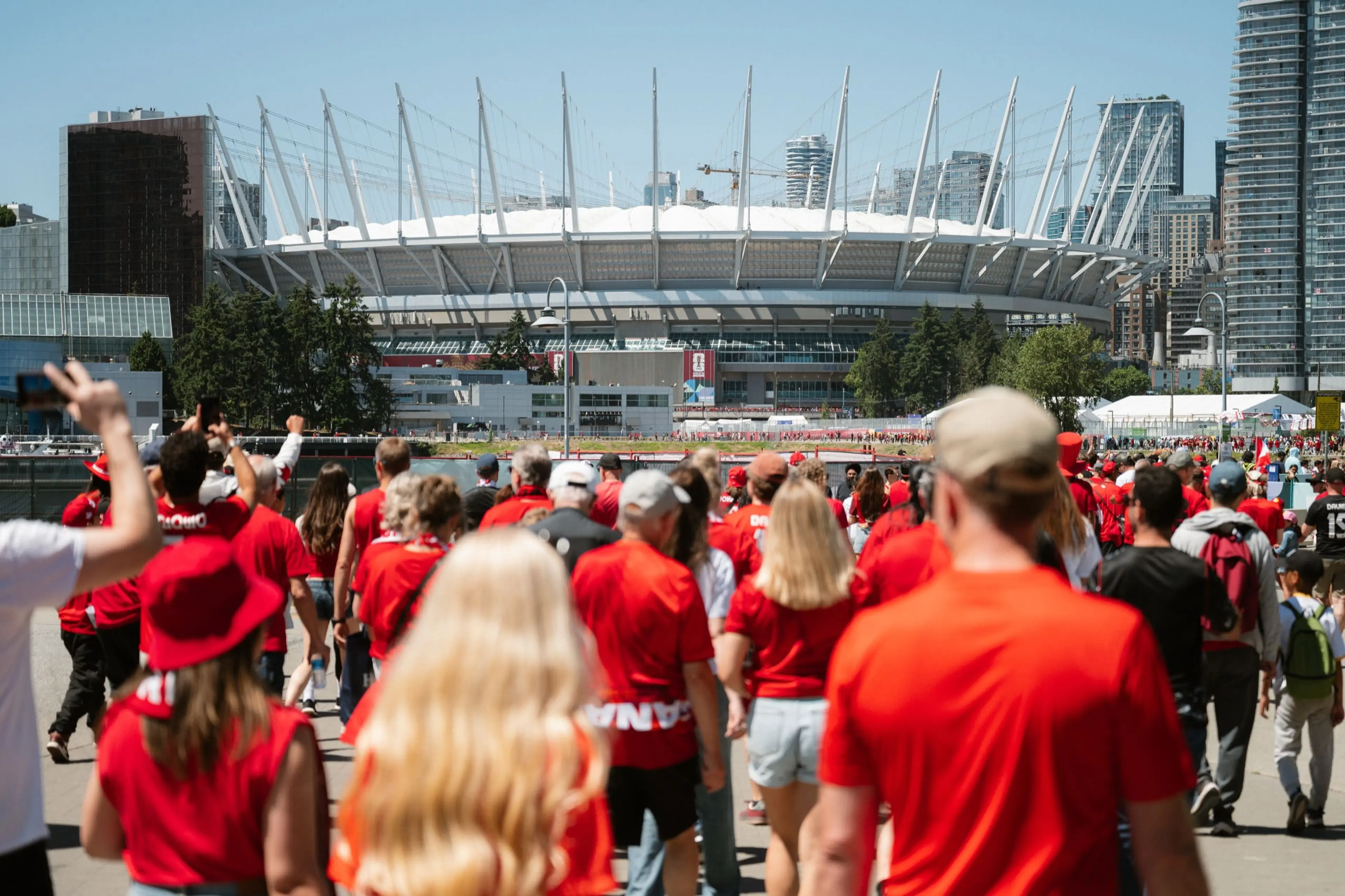 Crowd of people wearing red walking toward a large stadium with white spires on a sunny day.