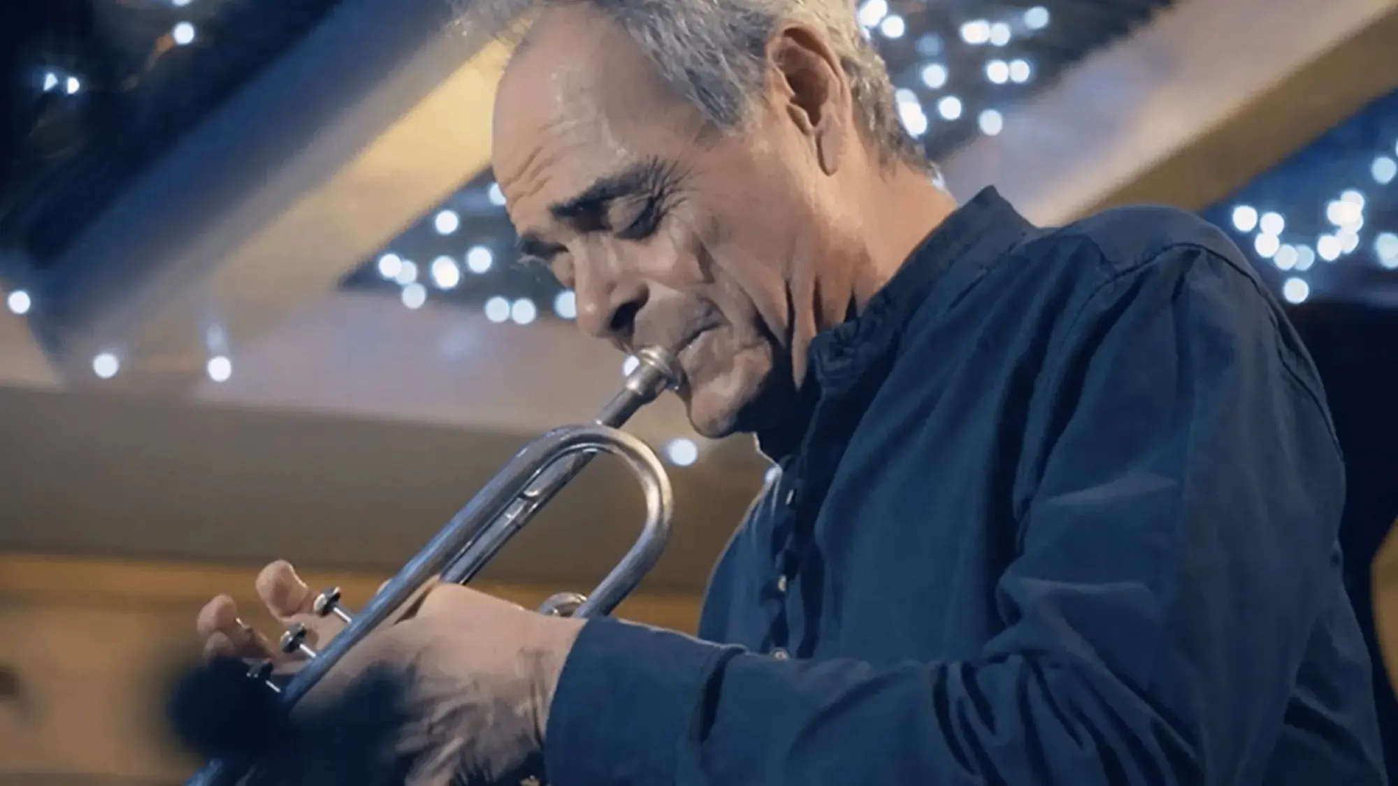 Person playing a trumpet indoors with string lights in the background.