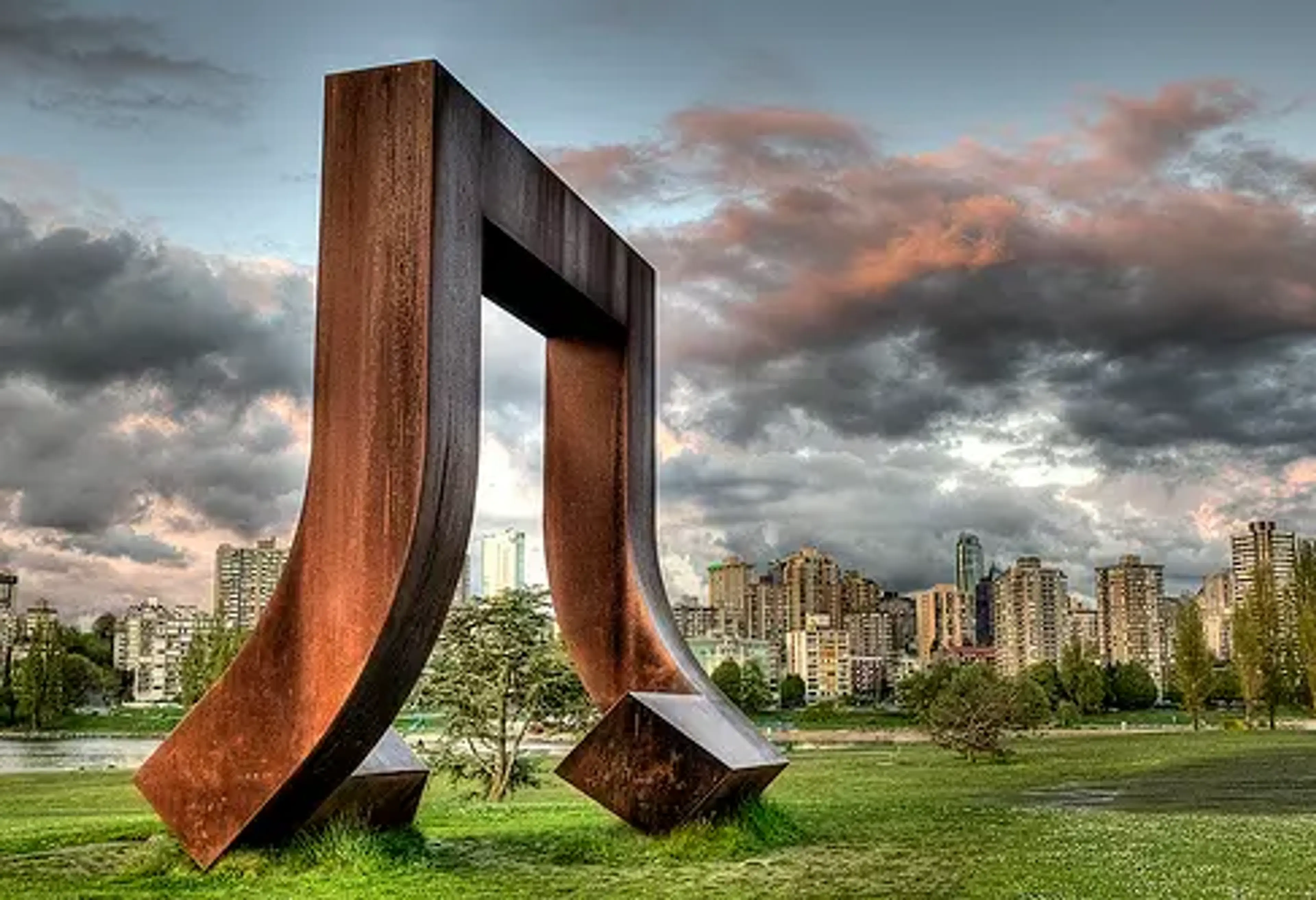 Large rust-colored abstract metal sculpture on grass at Vanier Park with city buildings and dramatic cloudy sky in background.