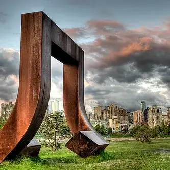 Large rust-colored abstract metal sculpture on grass at Vanier Park with city buildings and dramatic cloudy sky in background.