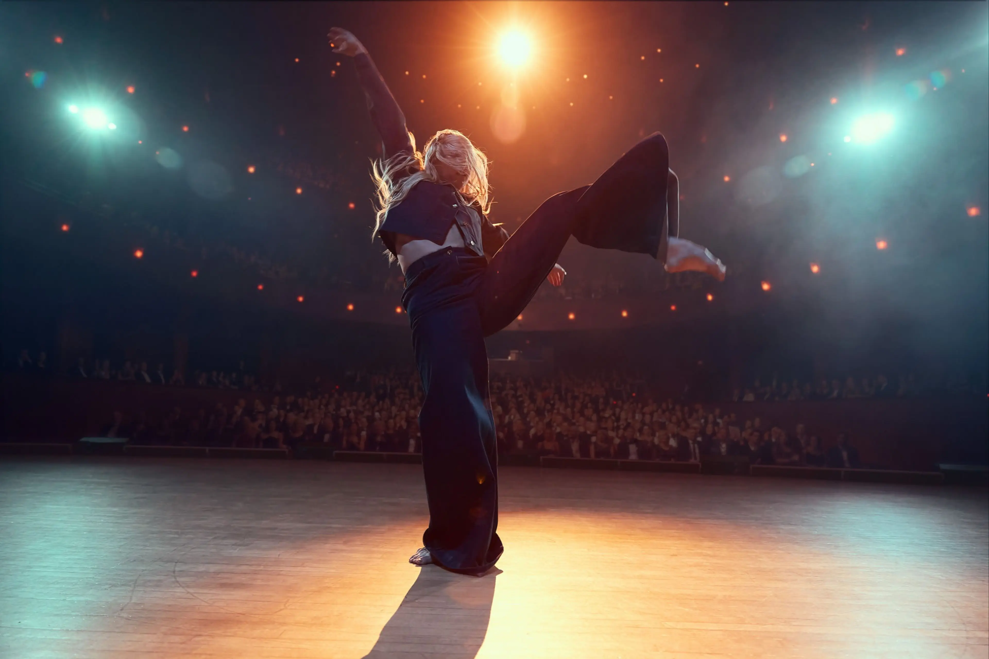 A person dancing on stage with one leg raised and arm extended, illuminated by stage lights with an audience in the background.