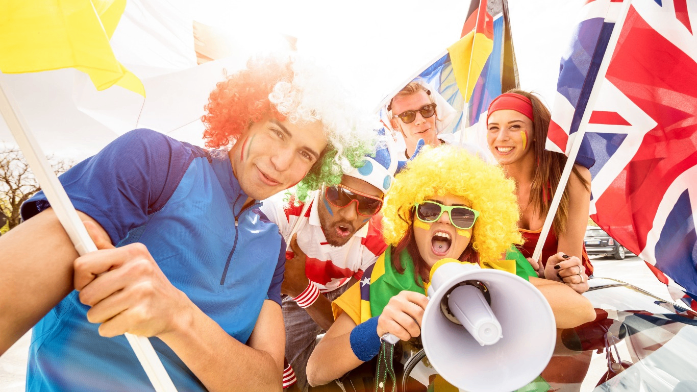 Five people wearing colorful wigs and face paint holding flags and a megaphone, cheering outdoors in bright sunlight.